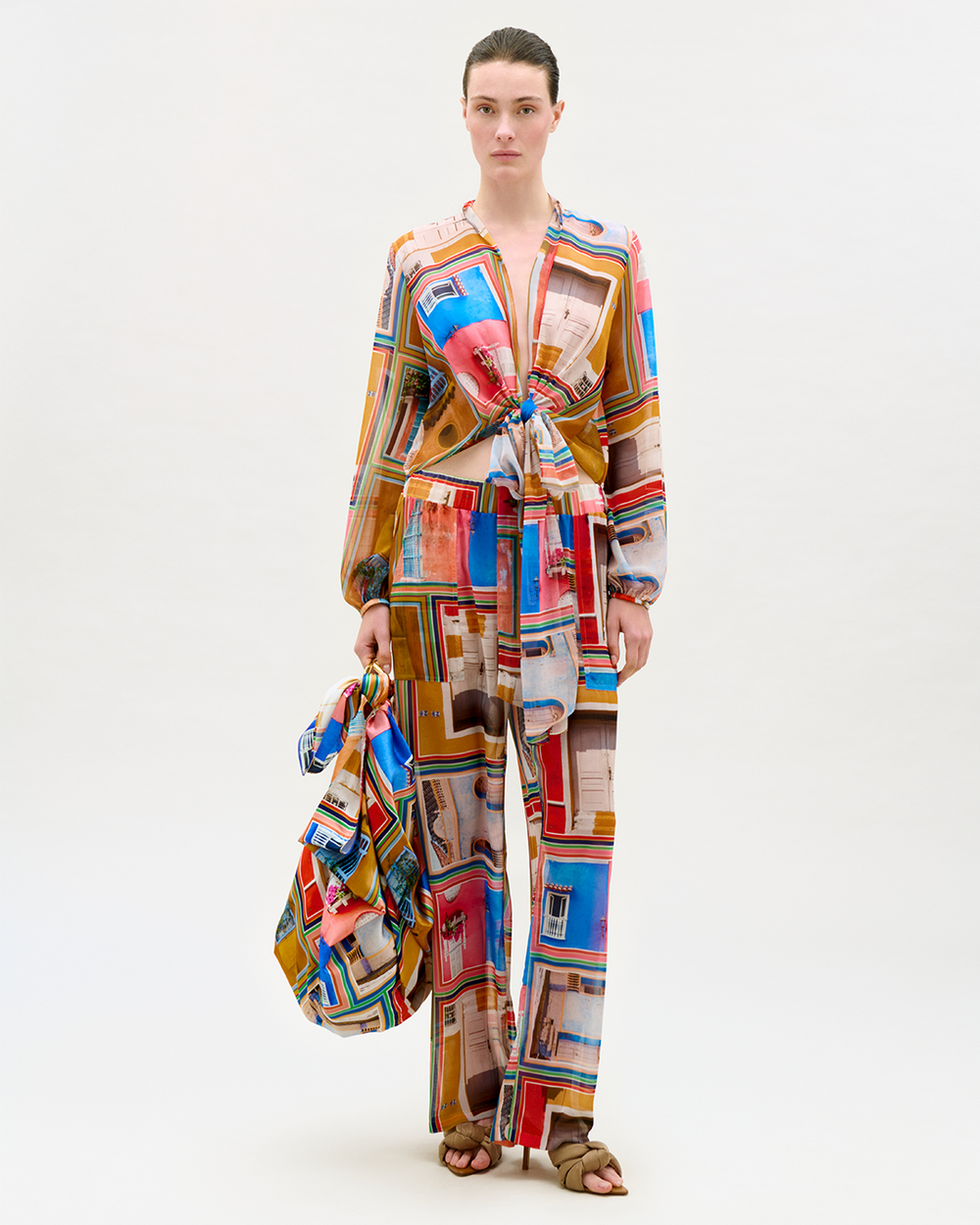 Model wearing a colorful long-sleeve blouse and wide-leg pants with abstract Cartagena-inspired graphics on a white background, holding a matching patterned bag