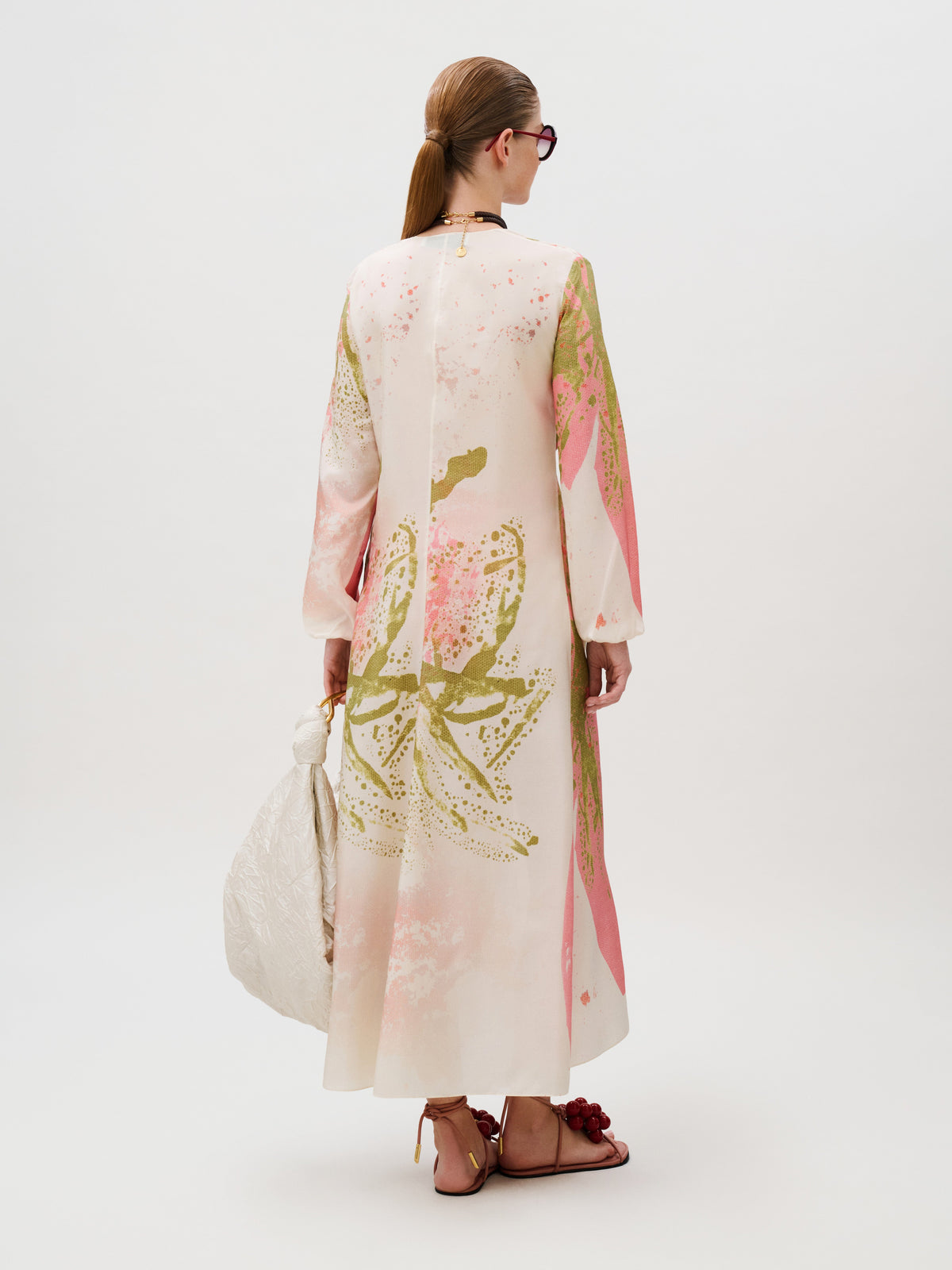 A person seen from behind wears a Cecelia Tunic in a cream-colored, long-sleeved silk kaftan with an abstract pink and green print, carrying a white gathered handbag and wearing strappy flat sandals.