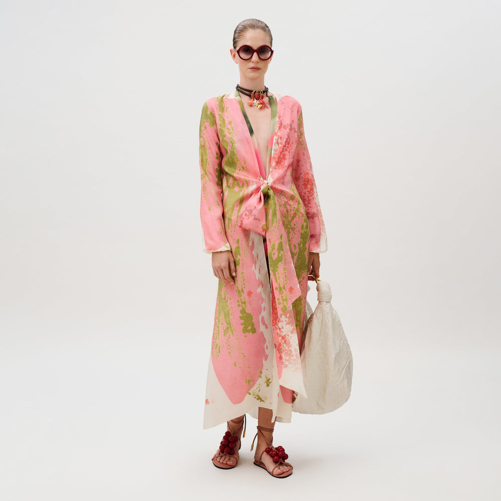 A model wears a knee-length, pink and green silk Cecelia Tunic with a front tie, dark sunglasses, beaded sandals, and a large white handbag.