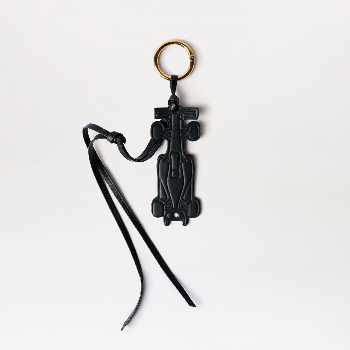 The F1 Charm is a black Formula 1 car-shaped keychain featuring a gold key ring and slim black leather straps, displayed against a white background.
