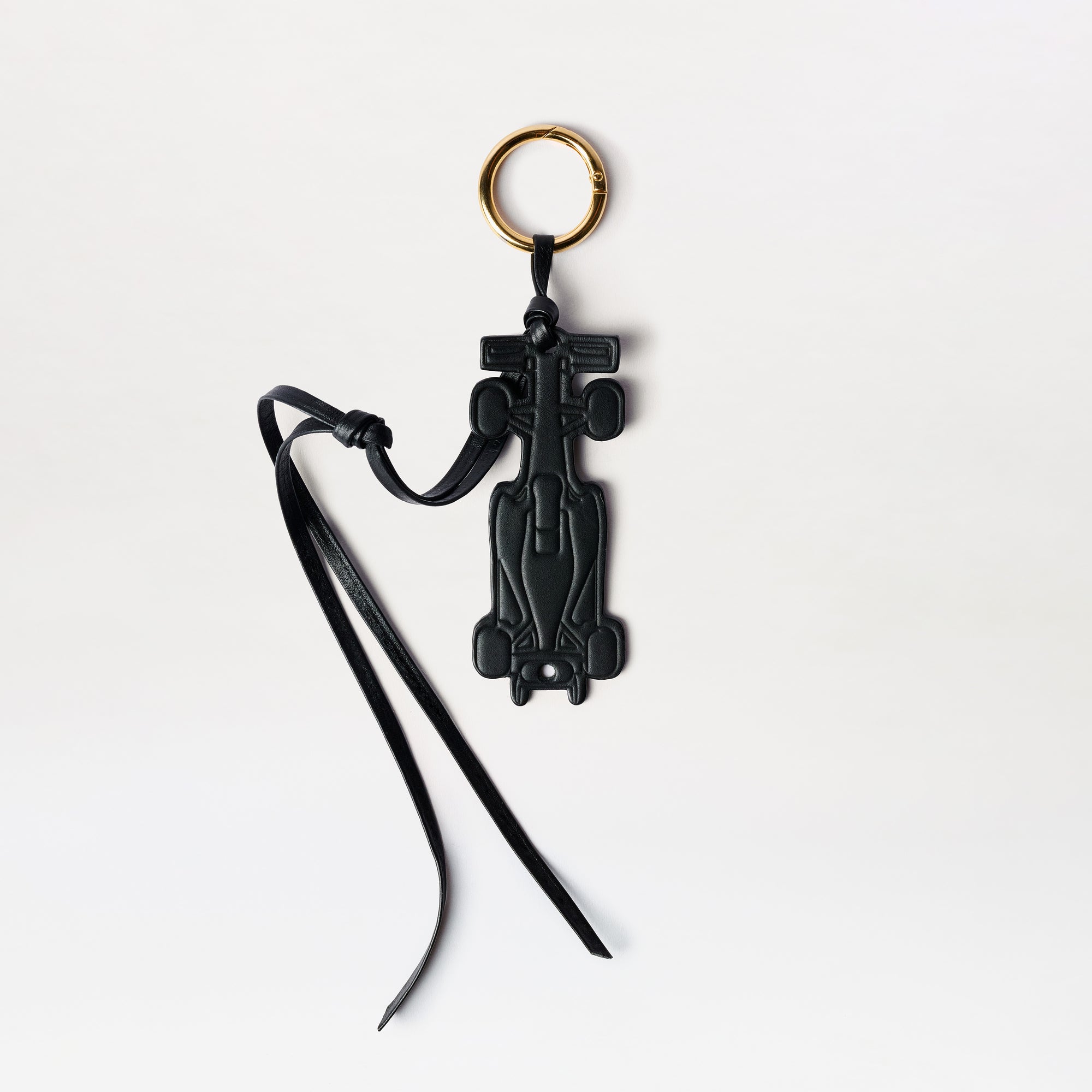 The F1 Charm is a black Formula 1 car-shaped keychain featuring a gold key ring and slim black leather straps, displayed against a white background.