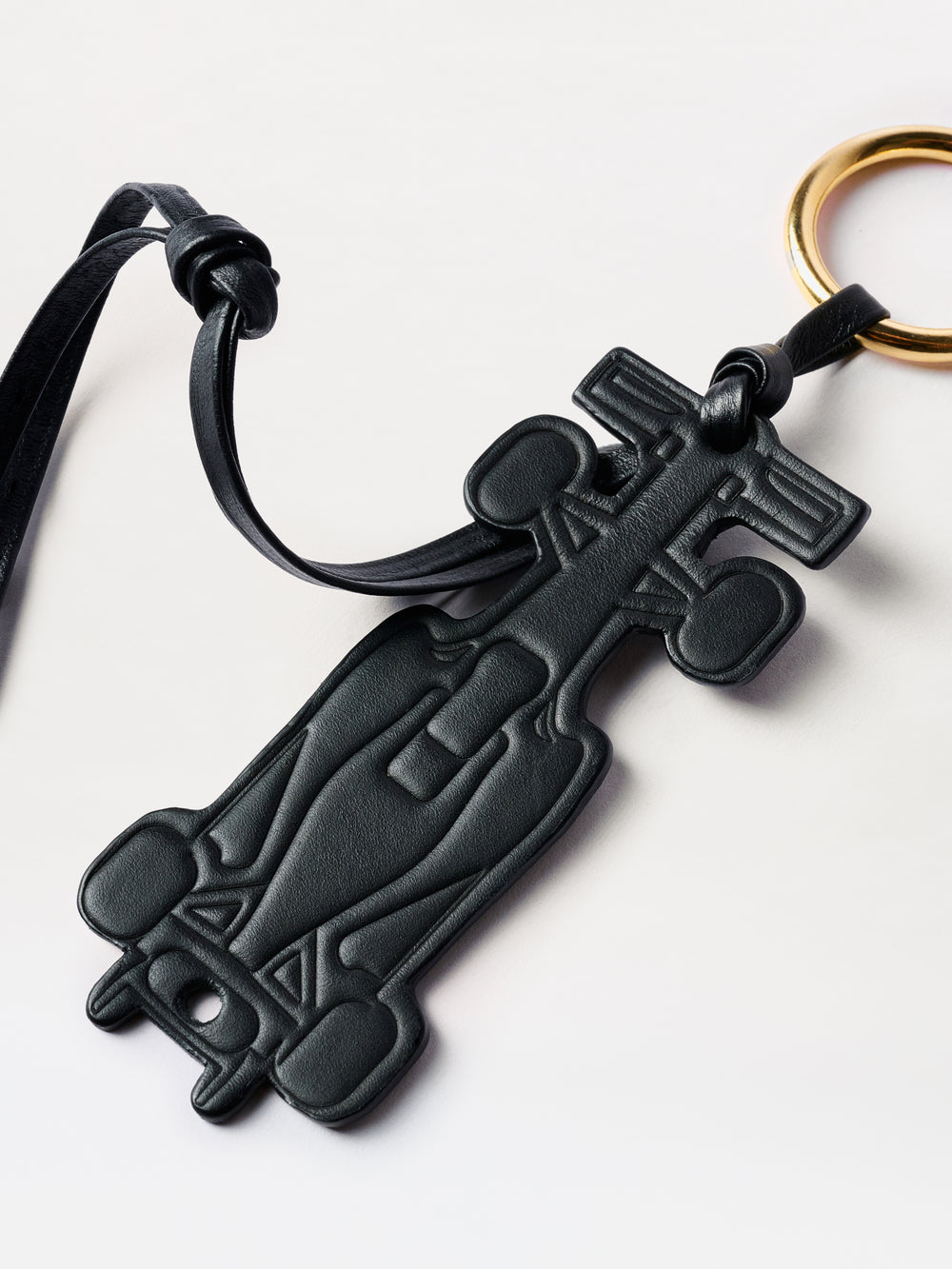 The F1 Charm is a black leather keychain in the shape of a Formula 1 race car, featuring a gold key ring and leather strap, displayed on a light background.