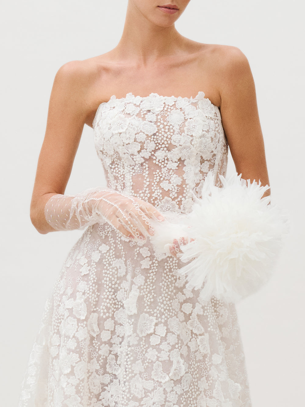 A woman wearing the Cindy Dress—a strapless white lace design featuring floral patterns—stands against a plain light background. Created by a Colombian atelier from luxurious Italian fabric, the dress is paired with sheer gloves and a fluffy white accessory. The image is cropped above her nose.