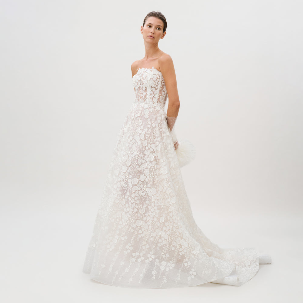 A woman wearing the Cindy Dress—a strapless, floor-length gown made from luxurious Italian fabric and adorned with intricate floral lace details—stands against a plain white background, gazing slightly upward with a calm expression.