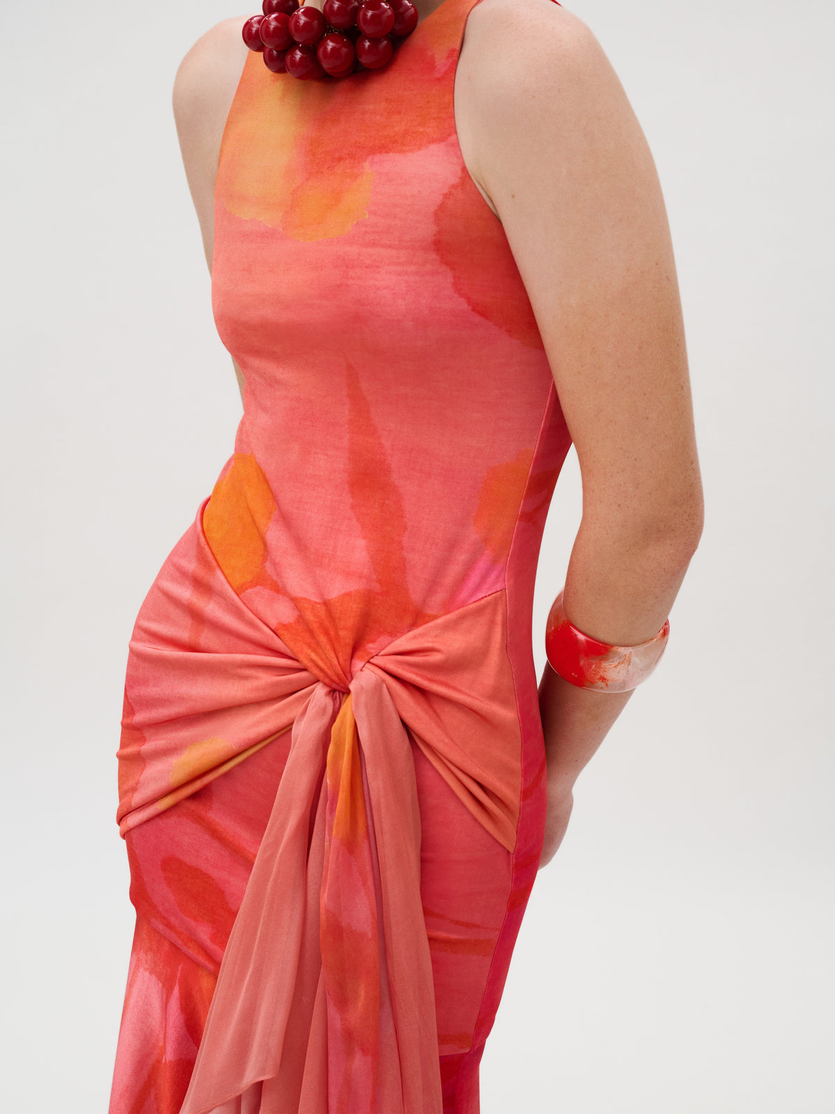 Clarice Dress, a slim-fit sleeveless midi dress in an Italian fabric jersey watercolor print in red and orange, featuring a gathered tie at the waist, worn with a large red bead necklace and a wide red bracelet.