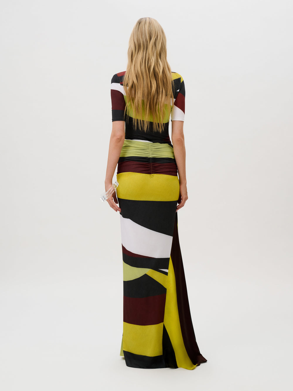 A person seen from behind wears the Claudia Skirt, a slim-fit, floor-length dress in Italian fabric with short sleeves, a multi-colored geometric pattern, and a gathered waist detail.