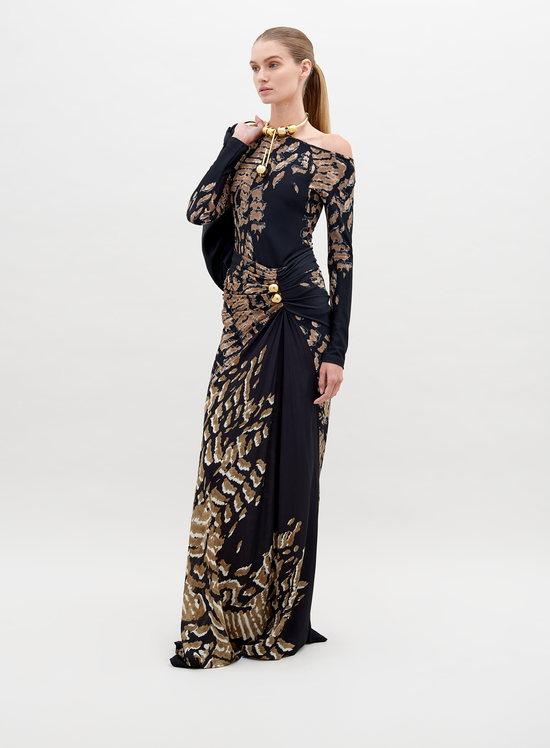 A woman stands wearing a long, fitted Claudia Skirt Black Abstract Animal Print. She has her hair pulled back and is accessorized with a gold necklace. Pre-order now to ensure delivery by the ship date of November 15th, 2024. The background is plain white.