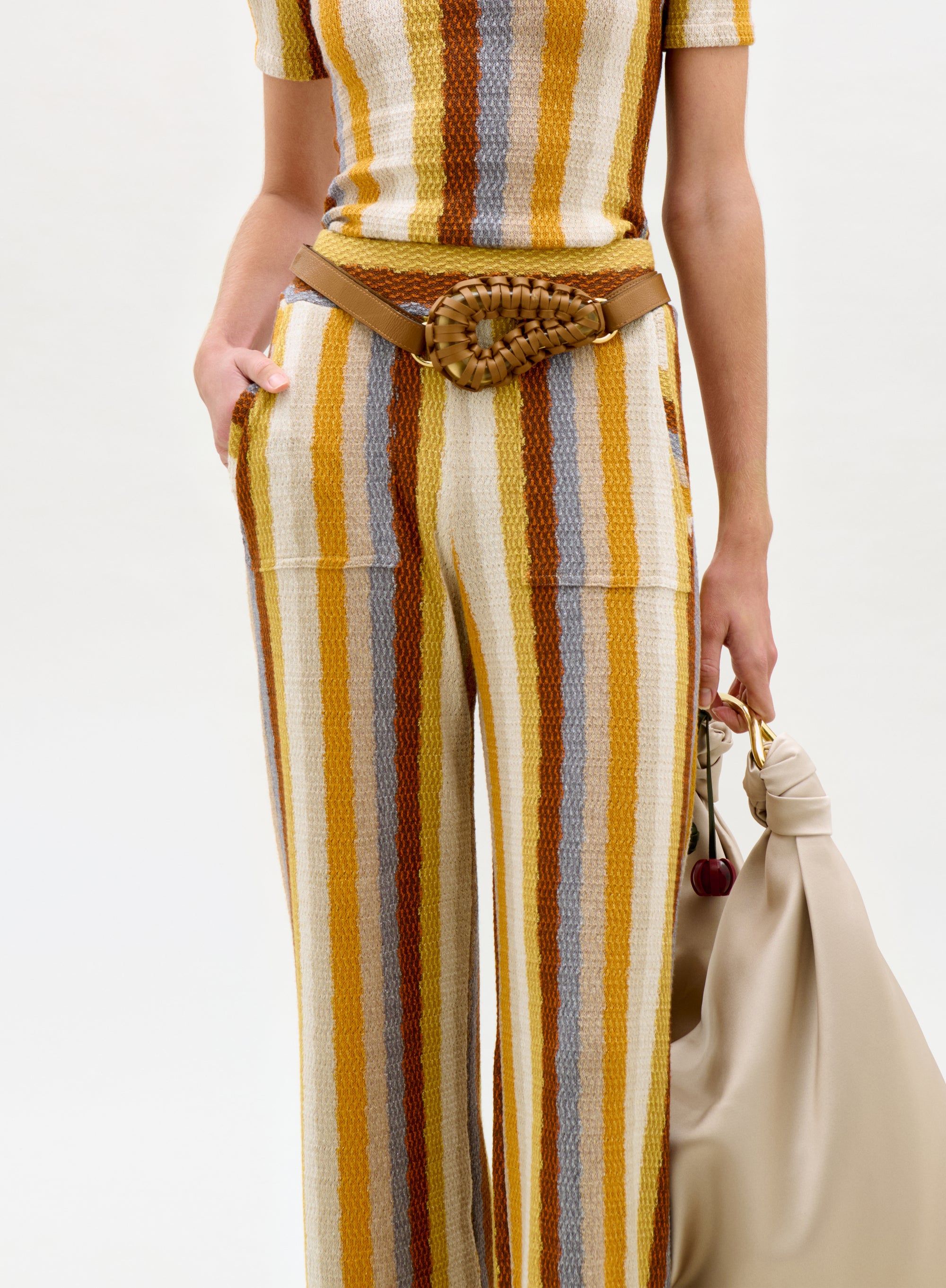 Person wearing a striped outfit with a belt and holding a beige bag on a white background