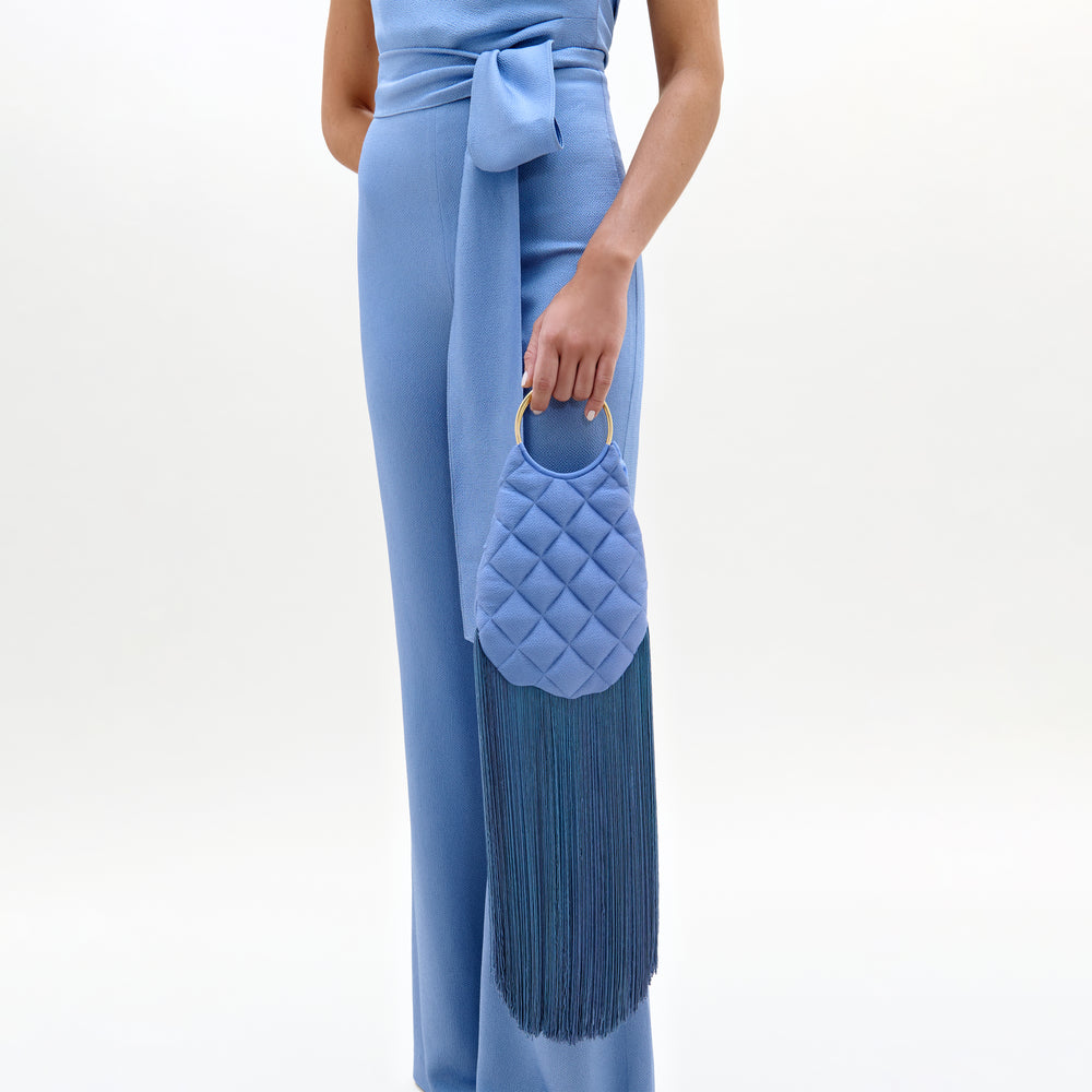 A person in a light blue jumpsuit with a sash holds the Toblach Bag, a structured oval Italian satin top-handle bag in light blue, featuring a gold ring handle and long blue fringe tassels. The background is plain white.