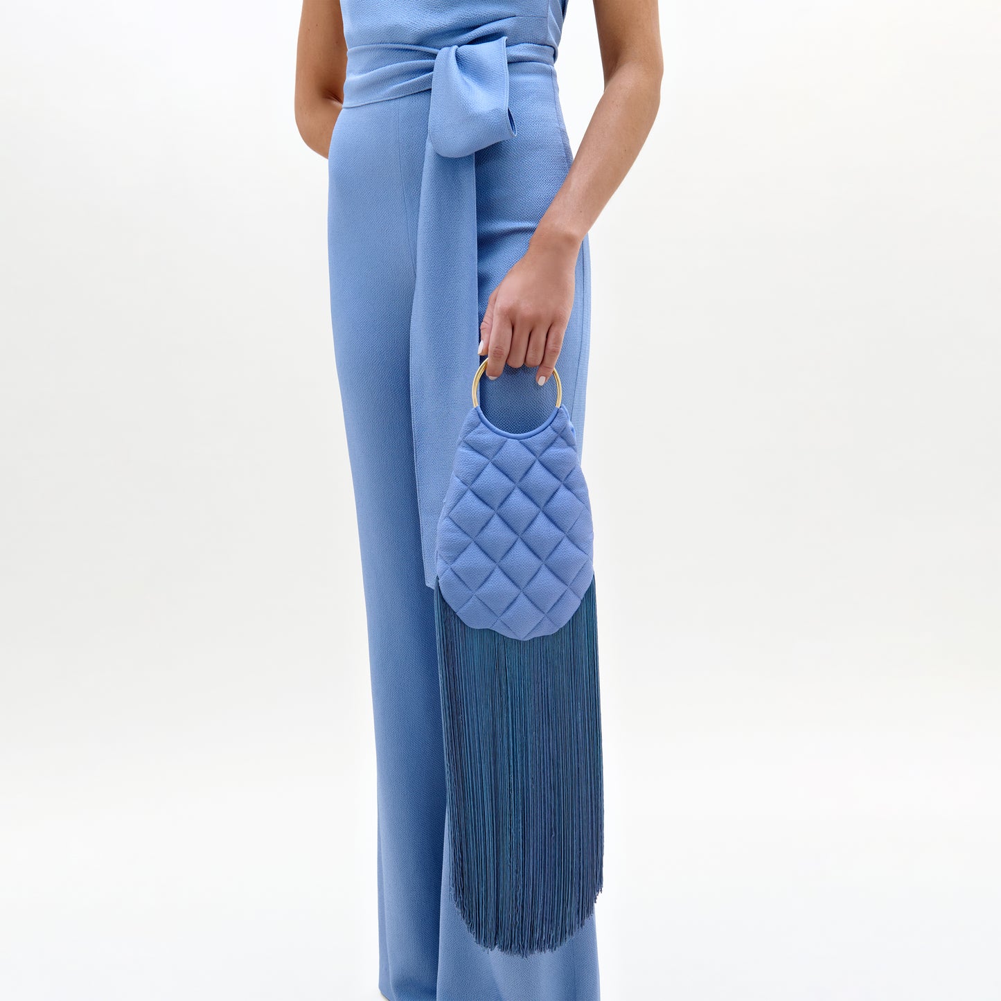 A person in a light blue jumpsuit with a sash holds the Toblach Bag, a structured oval Italian satin top-handle bag in light blue, featuring a gold ring handle and long blue fringe tassels. The background is plain white.