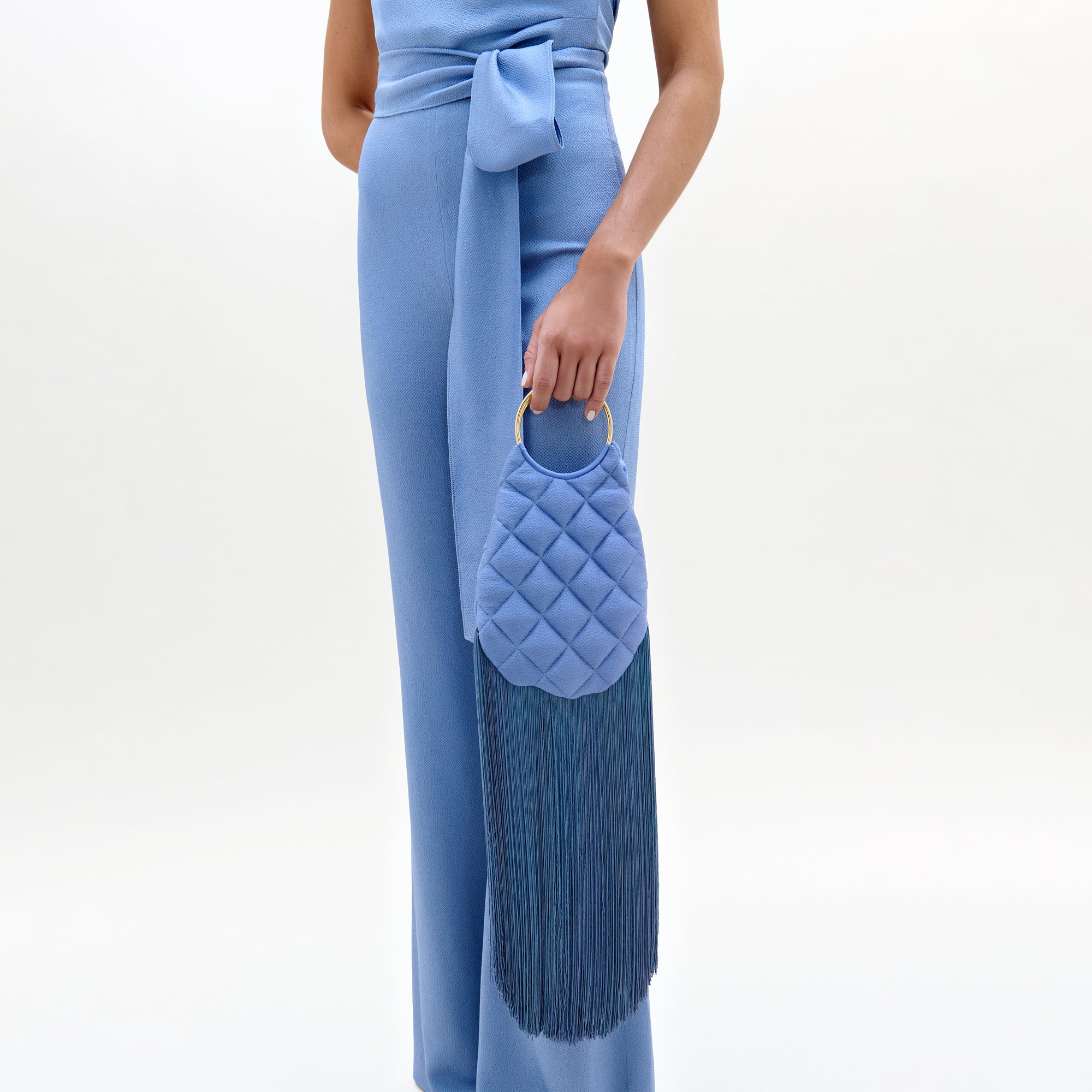 A person wears the Palermo Pant, a light blue sleeveless jumpsuit with straight tailored legs and a waist tie, holding a small quilted blue handbag with a gold handle and long fringe. The background is plain white.