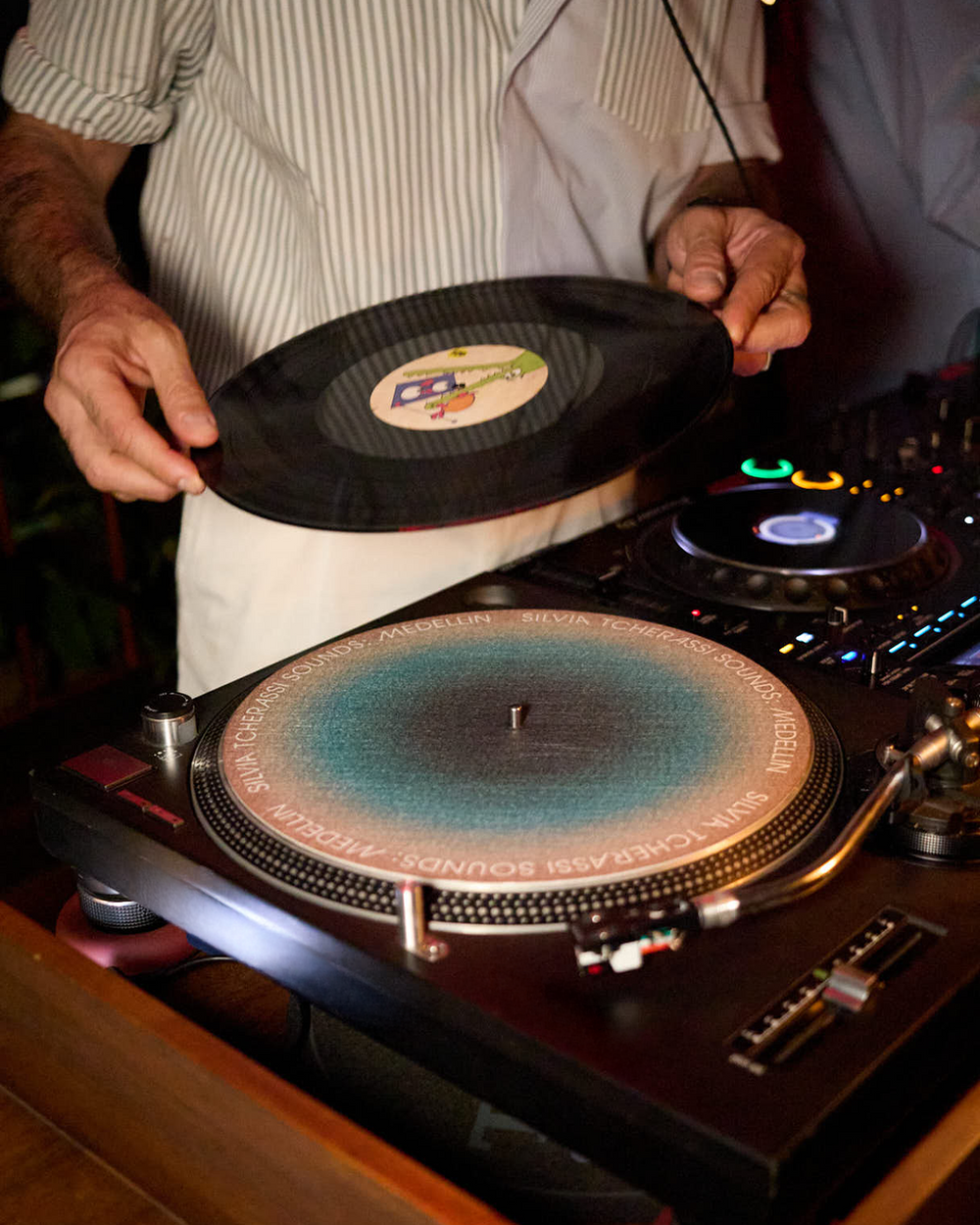 A DJ holding a black vinyl record above a turntable with a blue-to-pink gradient platter, in a dimly lit setting.