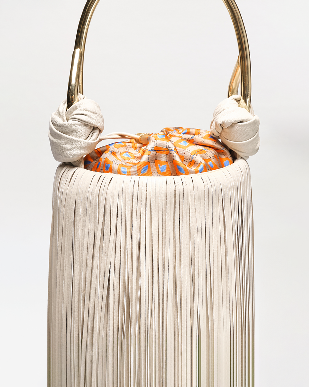 Handbag with white fringe, gold handle, and orange patterned interior on a light gray background