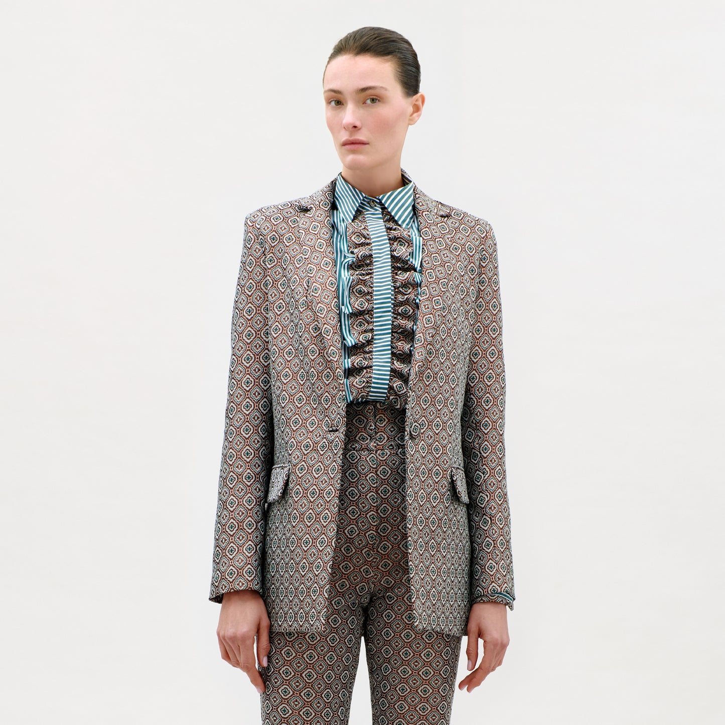 A person stands against a plain background wearing the Cuneo Jacket and matching trousers, expertly tailored from fine Italian fabric, with a striped, ruffled shirt. Their straight brown hair is pulled back, and they have a neutral expression.