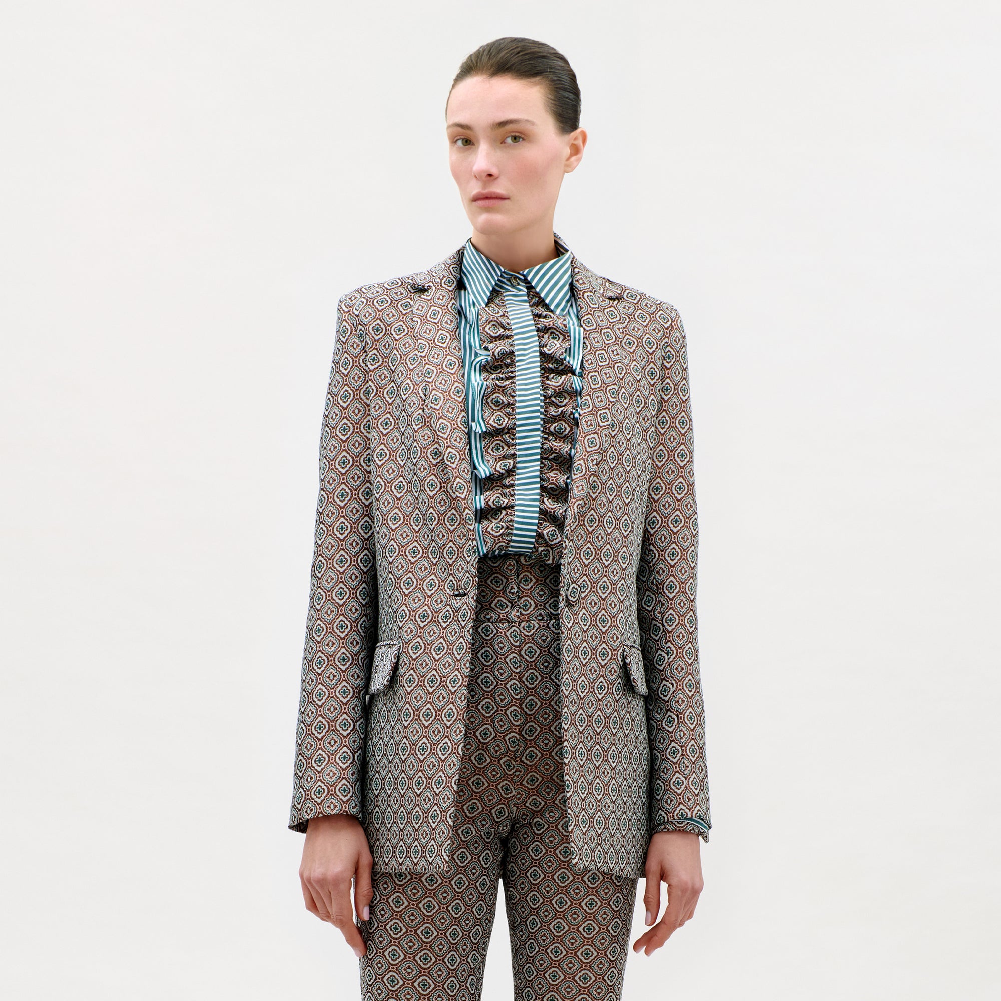 A person stands against a plain background wearing the Cuneo Jacket and matching trousers, expertly tailored from fine Italian fabric, with a striped, ruffled shirt. Their straight brown hair is pulled back, and they have a neutral expression.