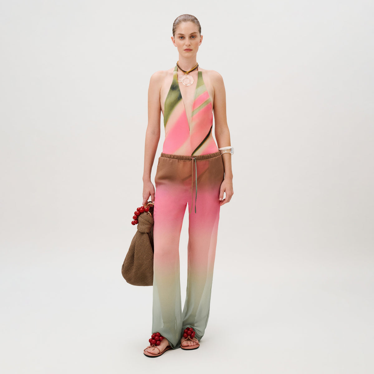 A model wears a pink and green gradient halter bodysuit and matching Curacao Pant, carrying a brown beaded tote bag and wearing matching beaded sandals.