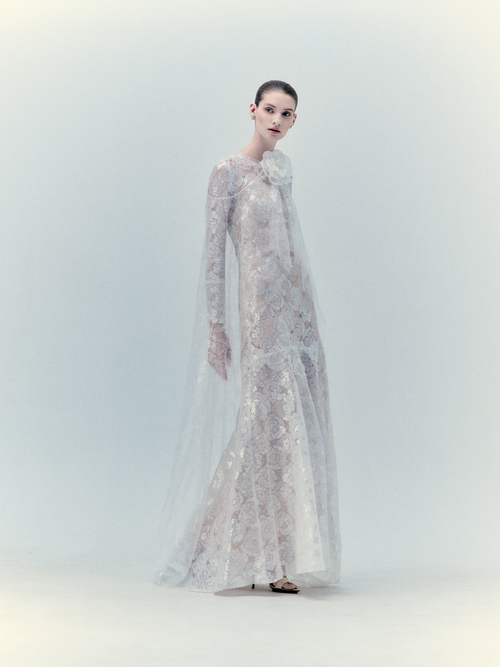 Person wearing the Curie Dress Pearl, a floor-length, long-sleeved, semi-transparent lace dress adorned with intricate floral patterns, standing against a plain, light-colored background. This exclusive piece is truly captivating; inquire for details.