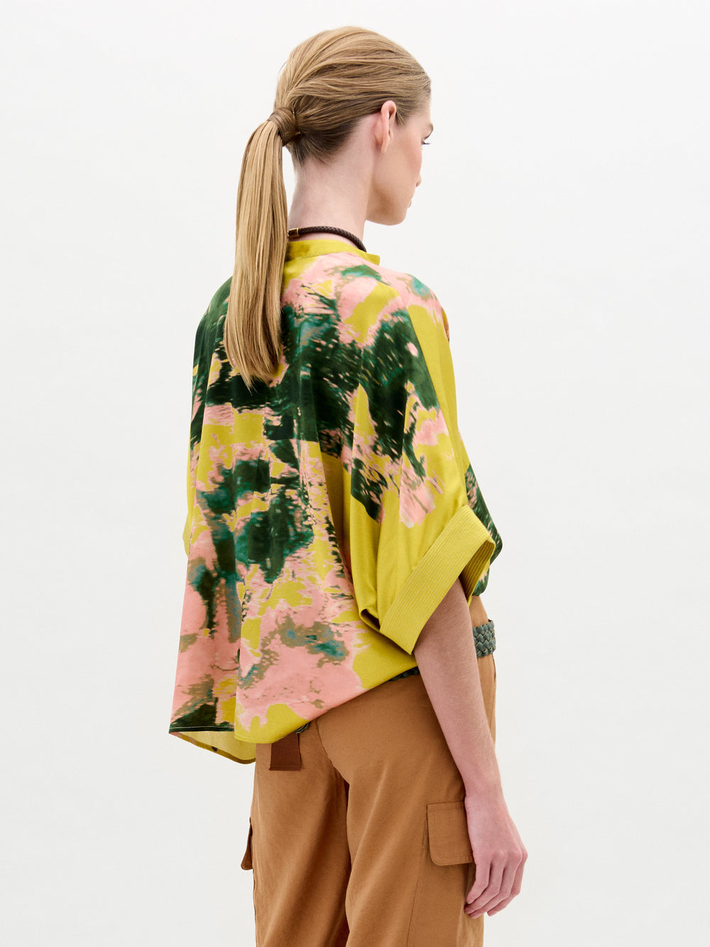 Woman wearing the Daia Blouse in a yellow floral print with an oversized fit, paired with tan pants, a green woven belt, and a choker necklace, standing against a plain white background.