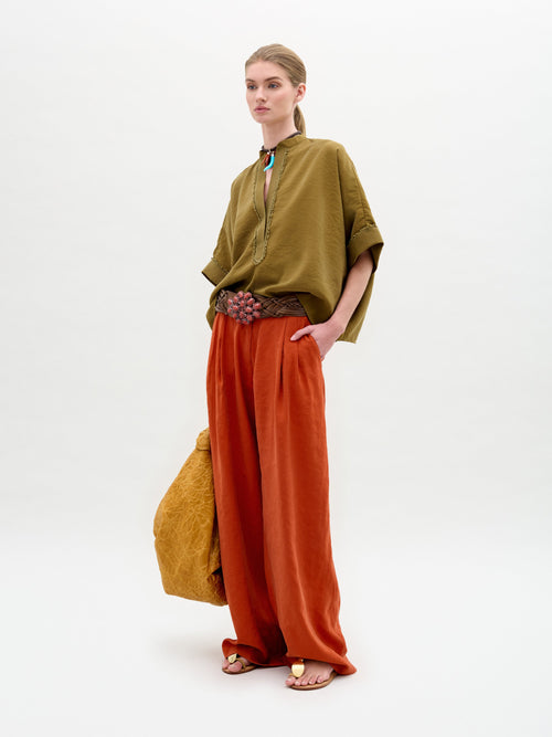 Wearing an oversized Daia Blouse in olive with a V-cut neckline paired with red wide-leg pants and sandals, they stand against a plain background holding a textured mustard-yellow bag made from exquisite Italian fabric.