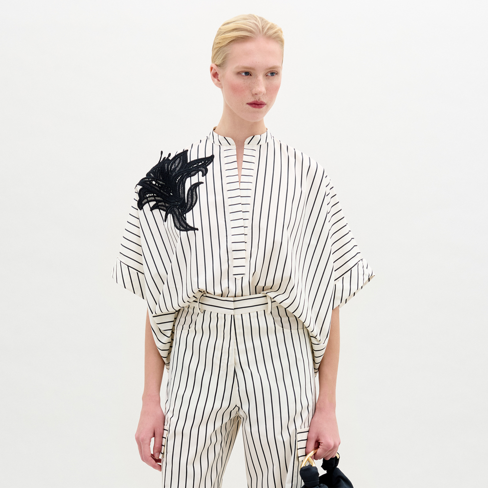 A woman in the Daia Blouse, a white short-sleeved, pinstriped top with a black abstract floral motif on the left shoulder and matching pants in Italian fabric, stands against a plain white background.