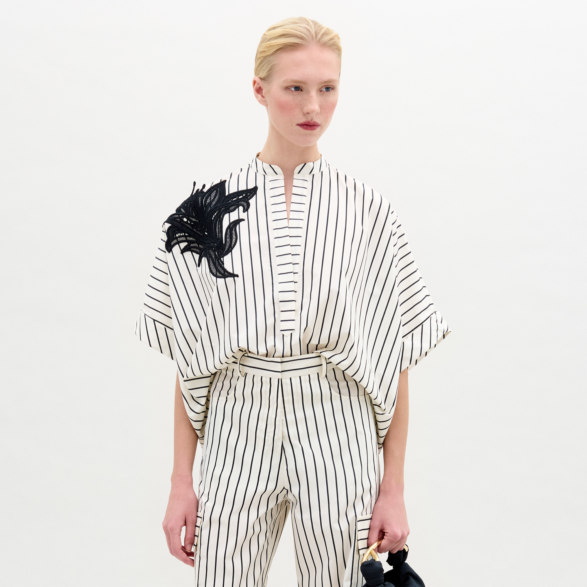 A woman in the Daia Blouse, a white short-sleeved, pinstriped top with a black abstract floral motif on the left shoulder and matching pants in Italian fabric, stands against a plain white background.