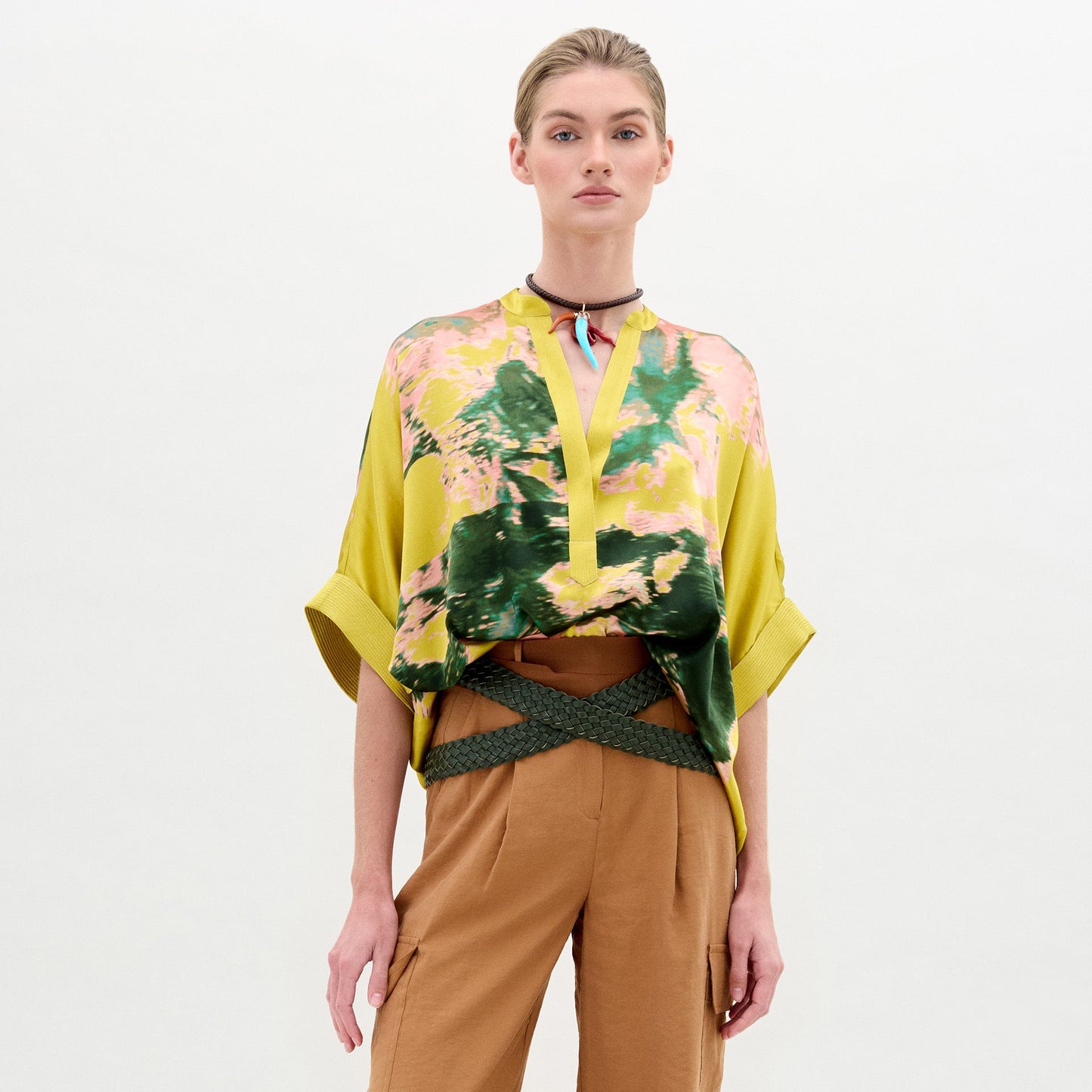 Woman wearing the Daia Blouse in a yellow floral print with an oversized fit, paired with tan pants, a green woven belt, and a choker necklace, standing against a plain white background.