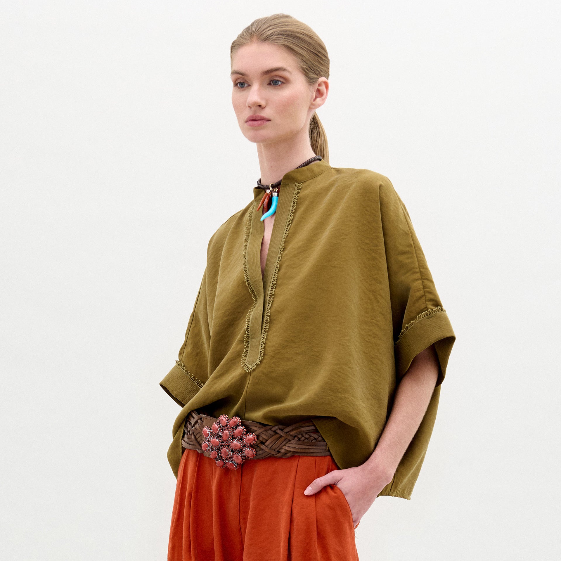 A woman stands sideways against a plain white background, wearing the Daia Blouse in olive green with a V-cut neckline, paired with loose orange pants and holding a textured yellow bag.