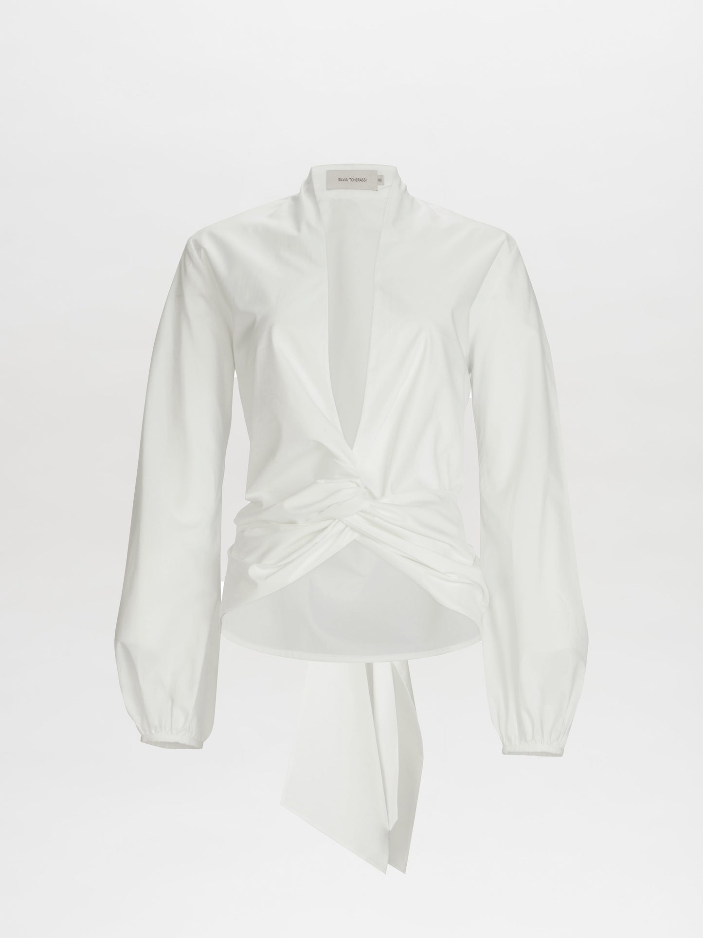 The Dania Blouse is a white long-sleeve top featuring a deep V-neck and twist knot detail at the front waist.