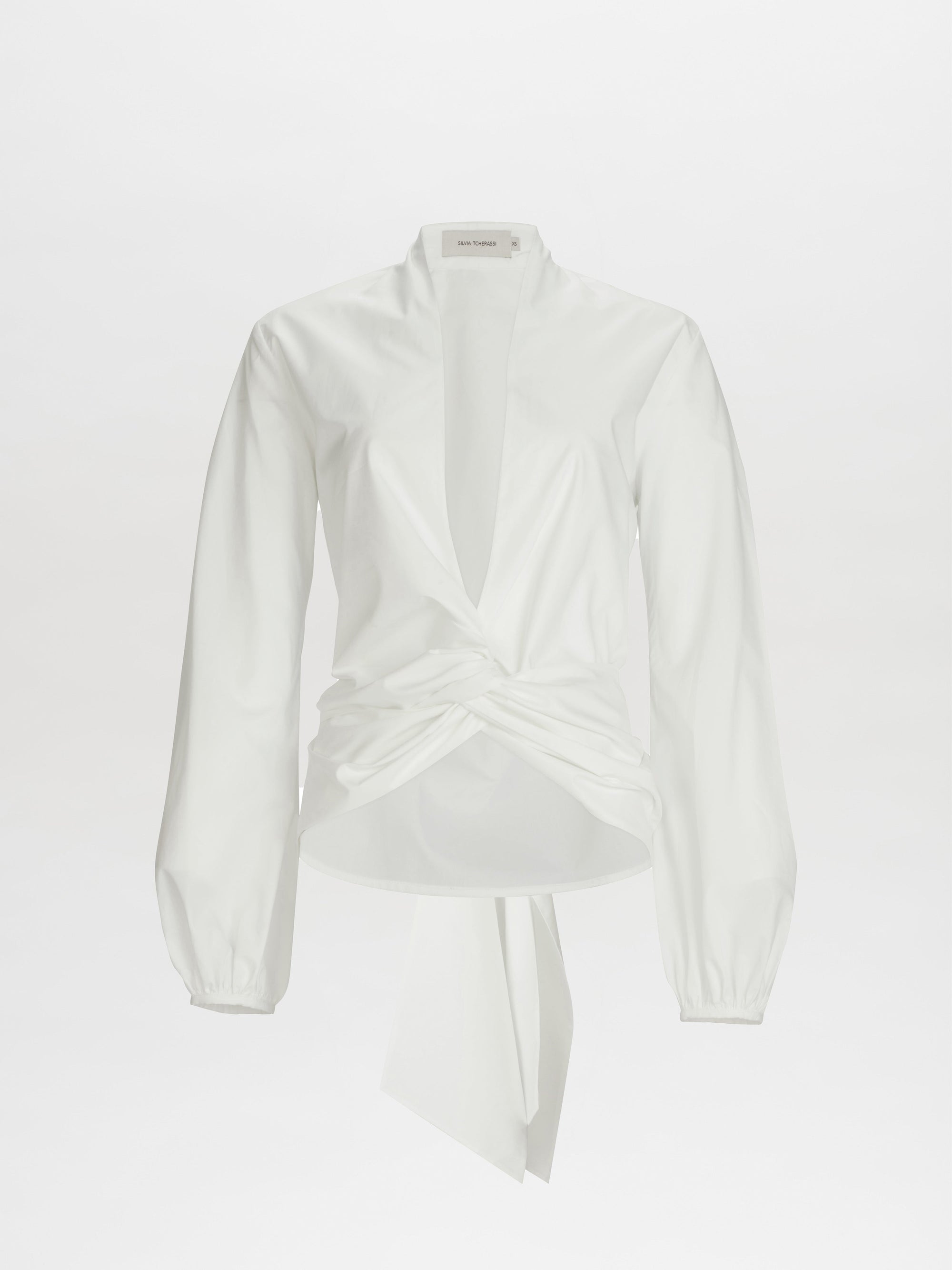 The Dania Blouse is a white long-sleeve top featuring a deep V-neck and twist knot detail at the front waist.