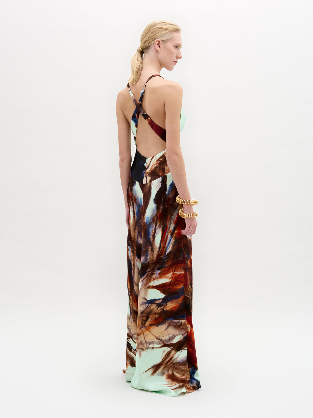 A woman stands against a plain white background wearing the Daniella Dress—a sleeveless maxi with a deep V-neckline, featuring a light blue top and a brown and red abstract print on the lower half.
