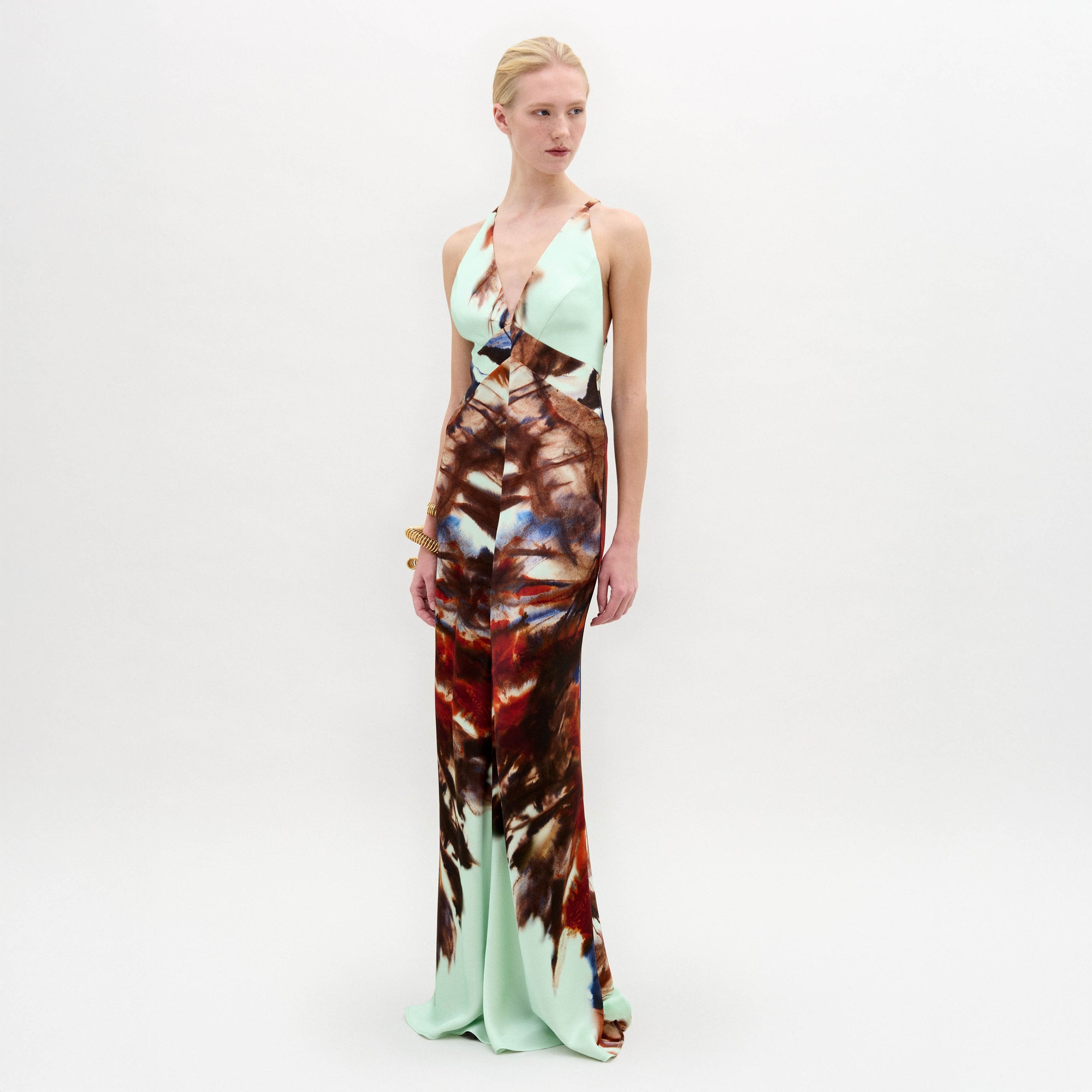 A woman stands against a plain white background wearing the Daniella Dress—a sleeveless maxi with a deep V-neckline, featuring a light blue top and a brown and red abstract print on the lower half.