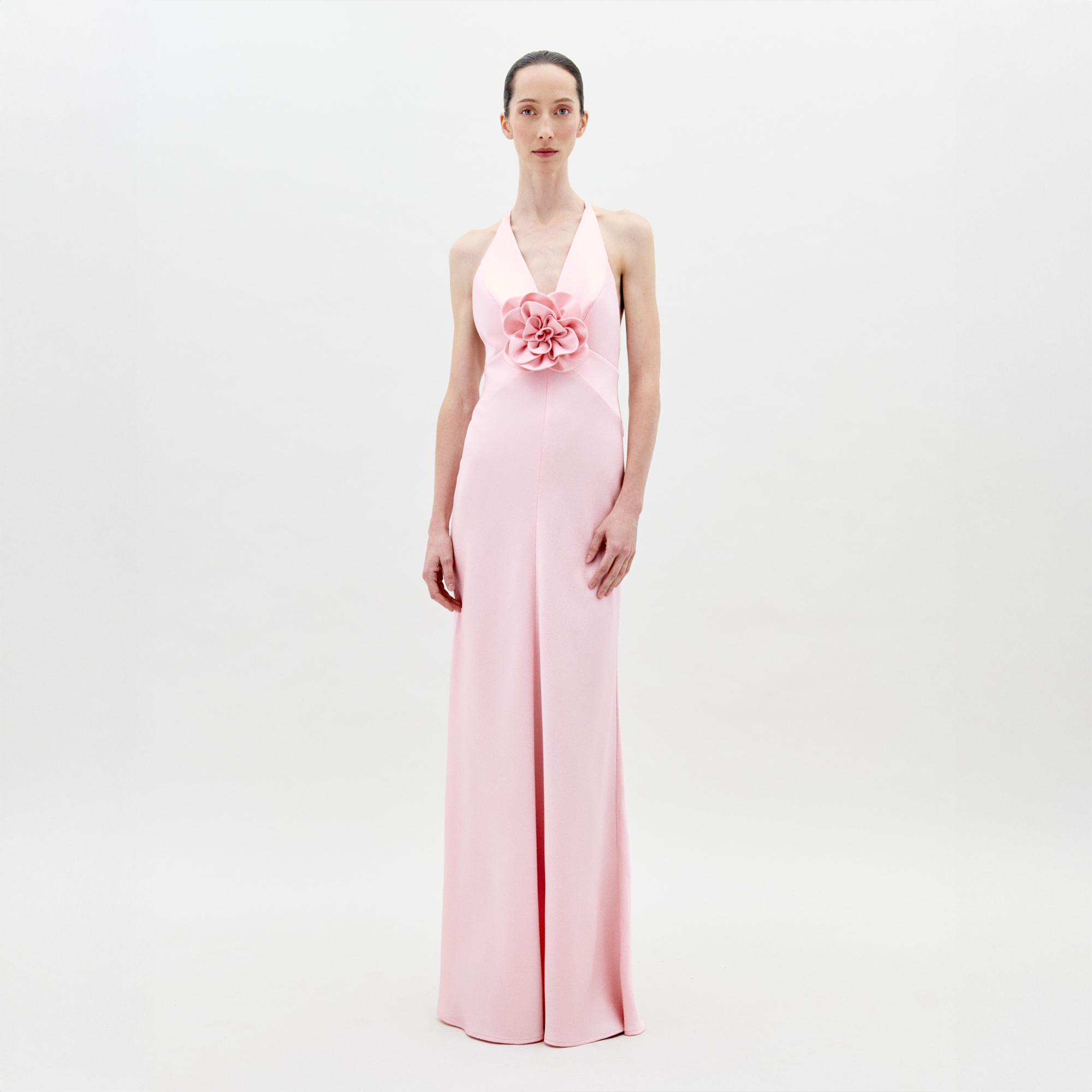 A person stands facing forward wearing the Daniela Dress Pink—a long, pastel pink dress featuring a halter neck and a large rose embellishment at the front. The background is plain white. Available for pre-order now, the dress will ship by February 1st, 2025.