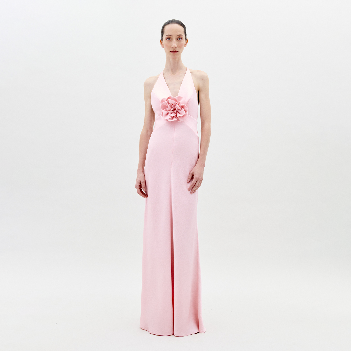 A person stands facing forward wearing the Daniela Dress Pink—a long, pastel pink dress featuring a halter neck and a large rose embellishment at the front. The background is plain white. Available for pre-order now, the dress will ship by February 1st, 2025.