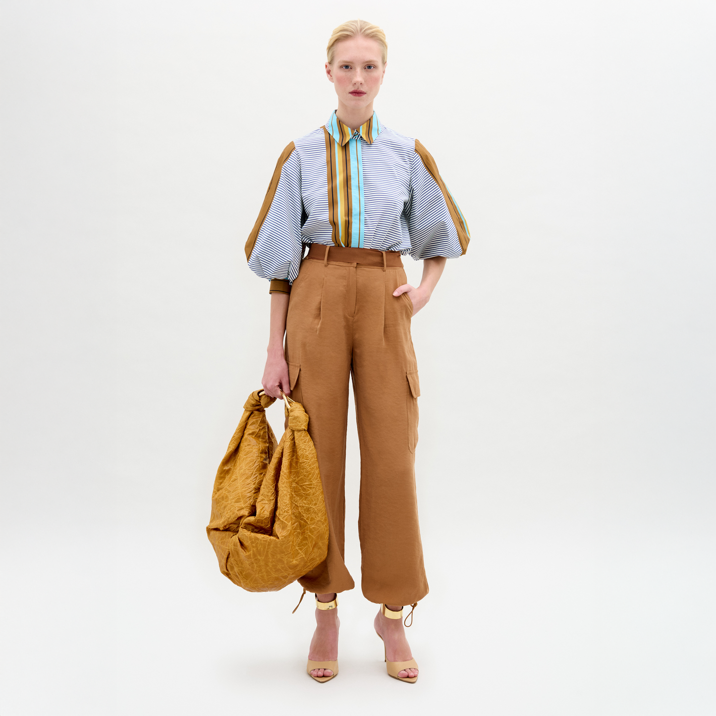 A woman stands against a white background wearing the relaxed fit, high-waisted Darnell Pant in tan cargo style, paired with a striped puff-sleeve blouse, open-toe heels, and holding a large tan bag.