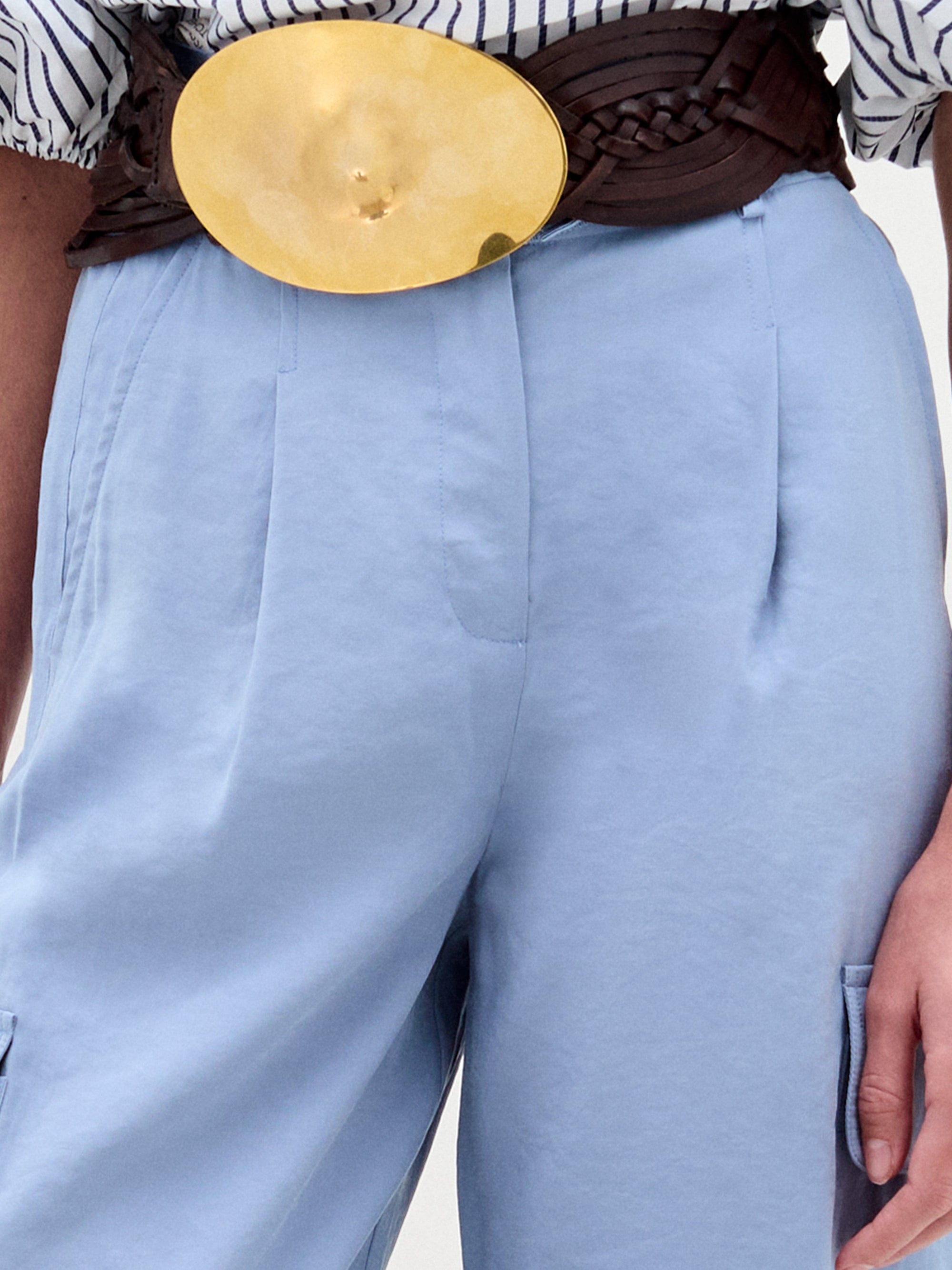 A woman stands against a plain background wearing a striped blouse with blue floral appliqué, the Darnell Pant in light blue, a brown belt with a large buckle, gold sandals, and holding a beige bag.