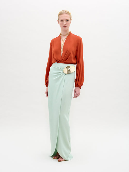 A person wearing an orange blouse and the Gadea Skirt in light green, featuring a large belt buckle, stands against a plain white background.