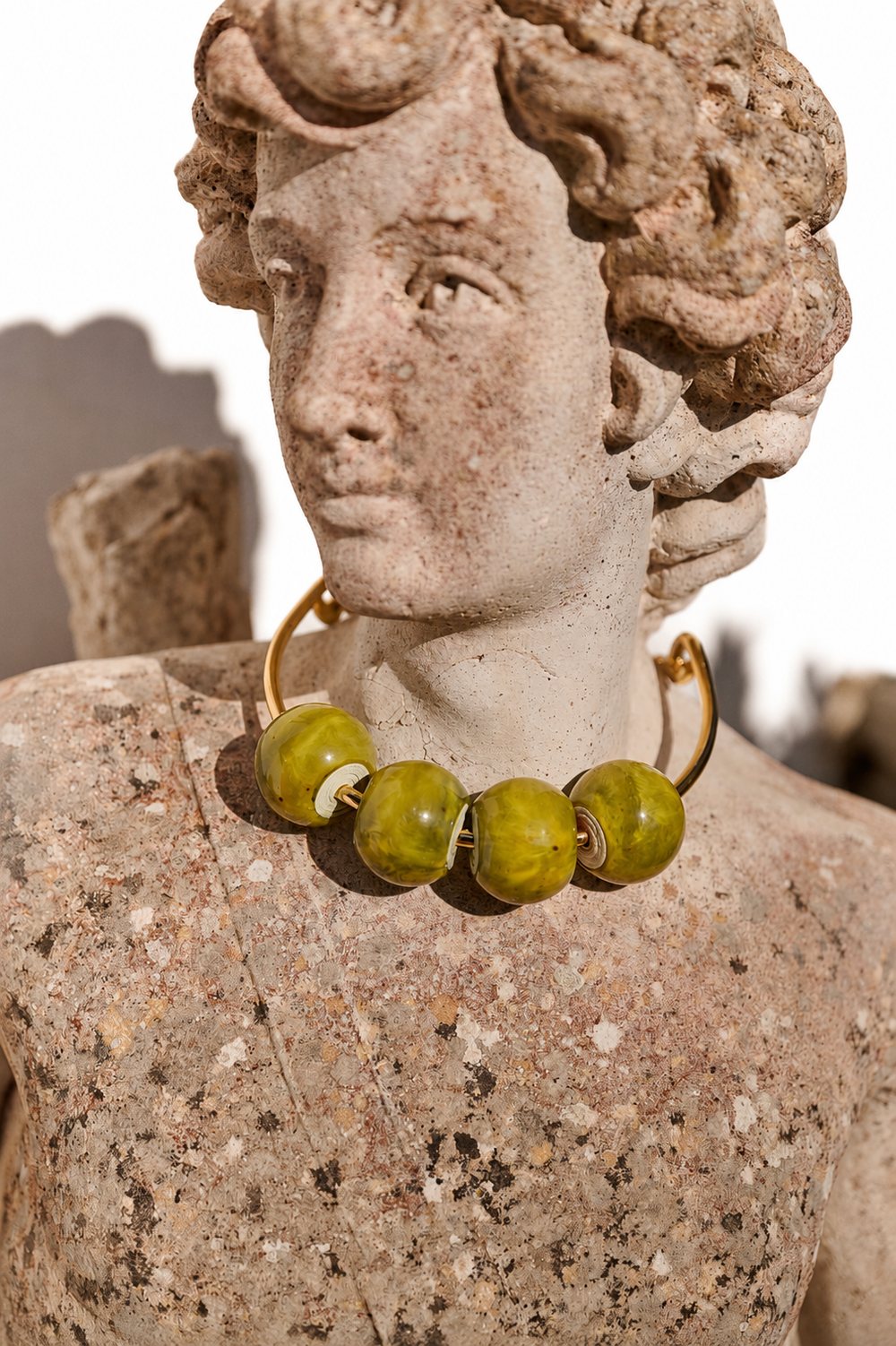 A close-up of a classical stone bust sculpture wearing a chunky gold-tone necklace with large olive-green beads, set against a bright white background.