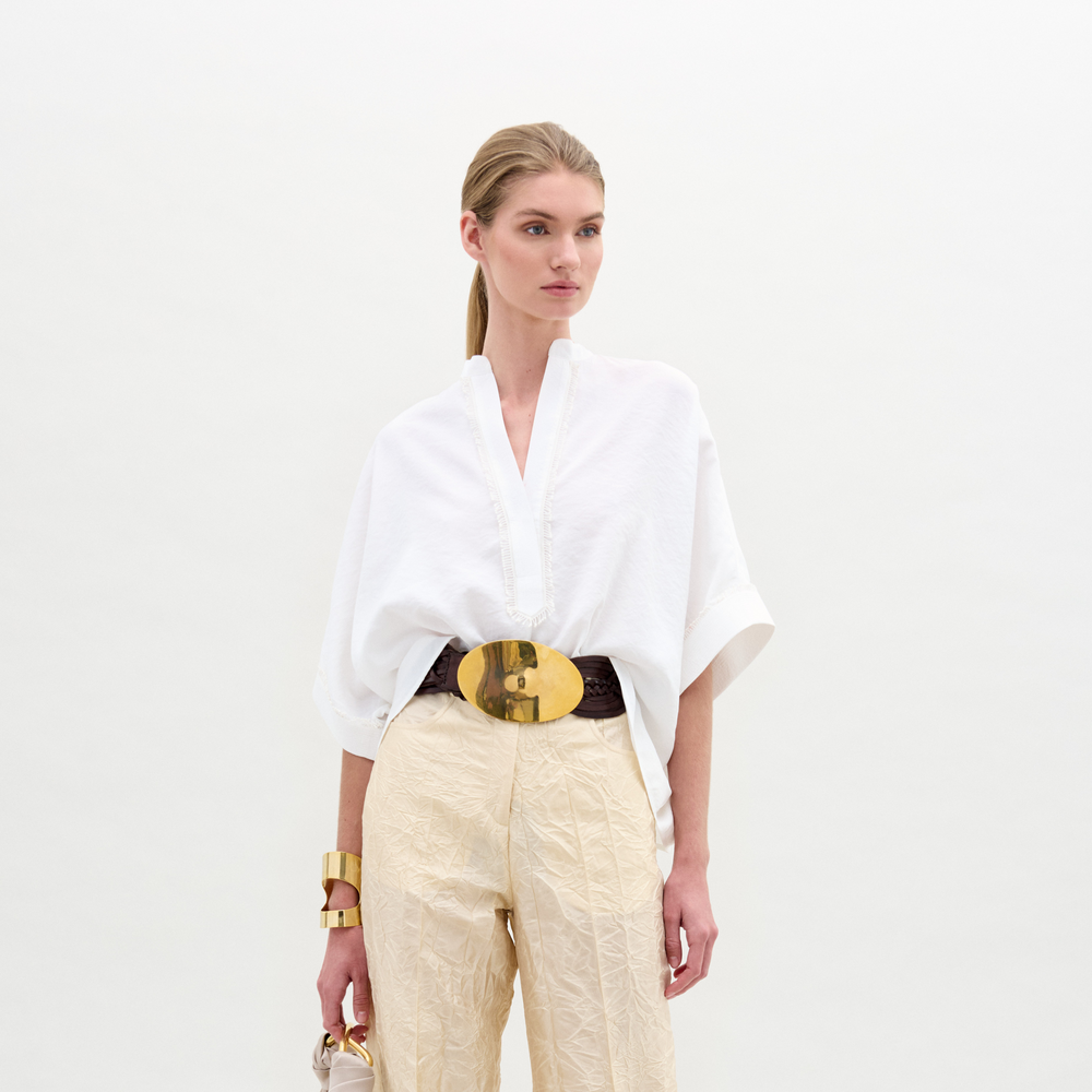A person in a Daia Blouse paired with beige pants and a large belt stands against a plain background, accessorized at the wrist.