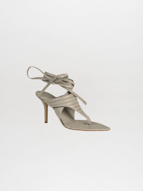 The Dalila Heels Grey are a stunning single strappy high-heeled sandal crafted from Italian calf leather in grey, featuring elegant wraparound ankle straps against a white background.