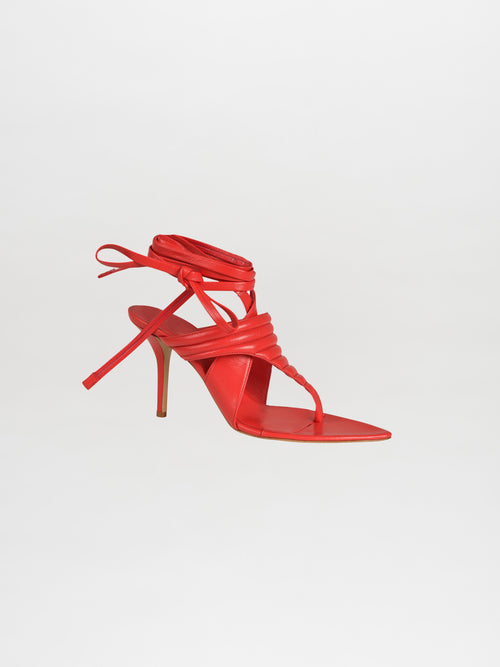 The Dalila Heels Red are a single red high-heeled sandal featuring wraparound ankle straps, highlighted against a pristine white background.