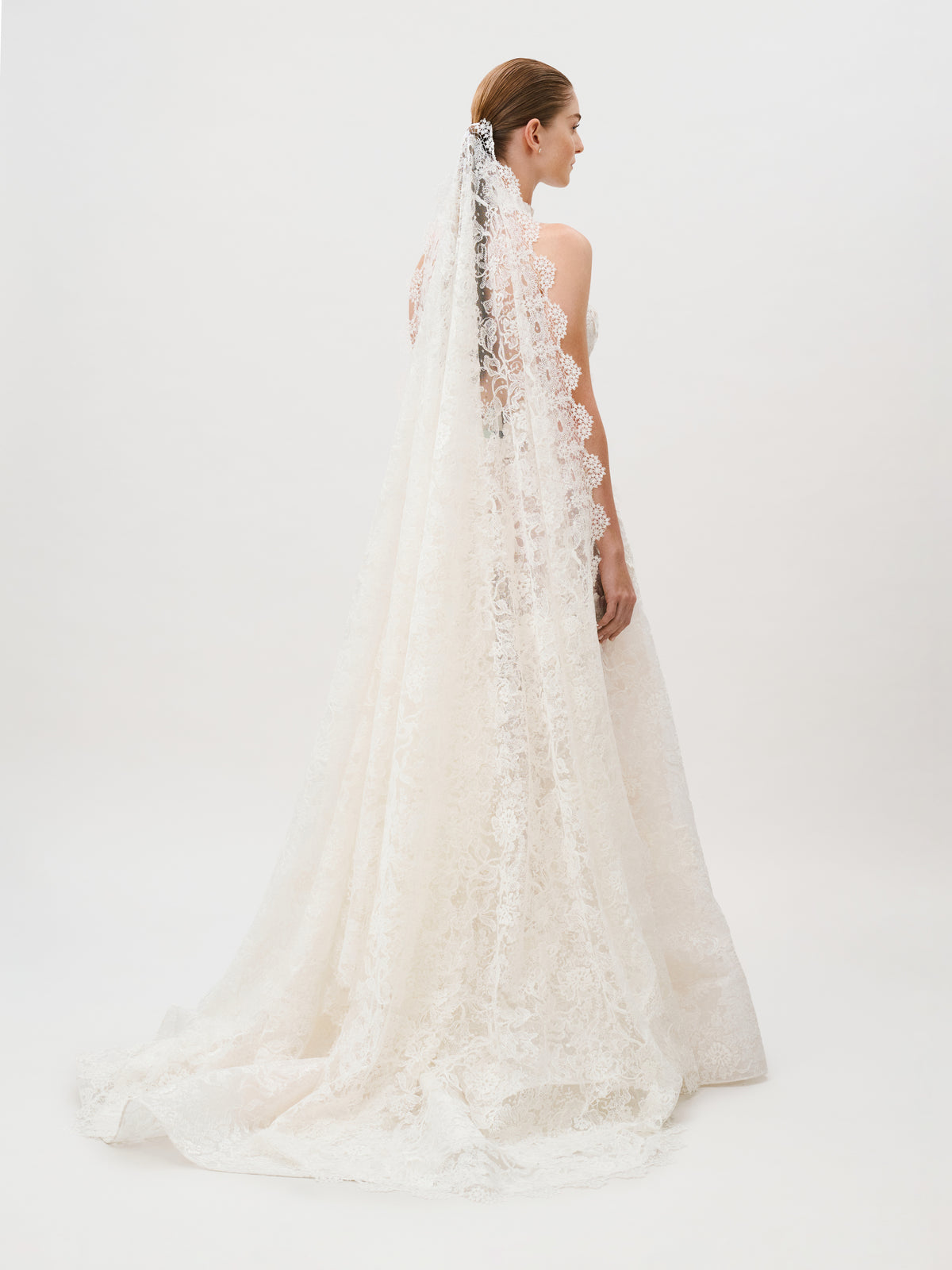 A bride in the Danita Dress—a white lace wedding gown crafted from luxurious Italian fabric—stands with her back to the camera, a long, elegant lace veil trailing behind her against a plain white background: a true vision of timeless beauty from Colombia Atelier.
