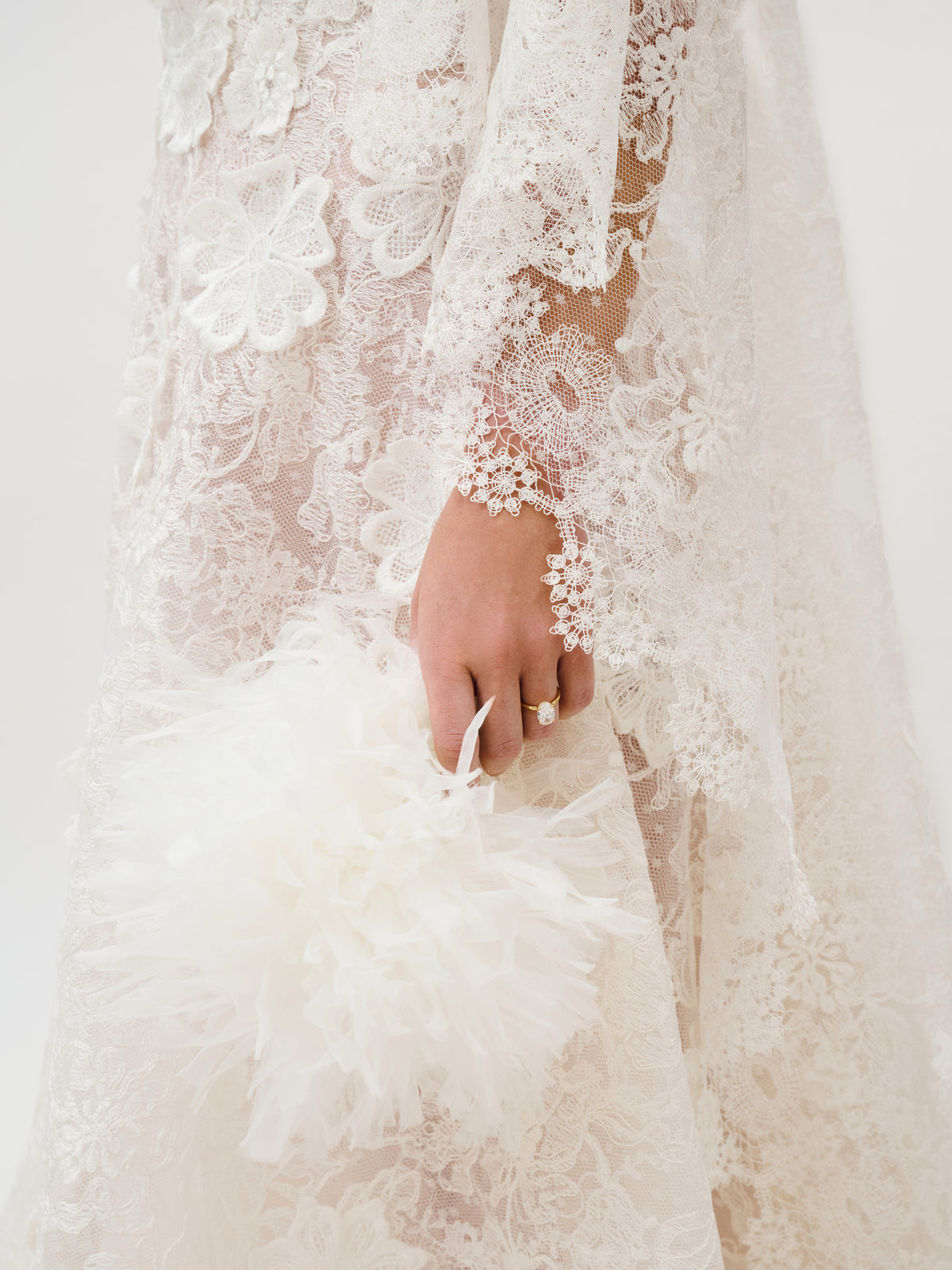 A person wearing the Danita Dress, made from exquisite Italian fabric by Colombia Atelier and adorned with intricate white floral lace, holds a fluffy white feathered accessory. Their hand displays a ring, while the light, neutral background complements the detailed elegance of the dress.