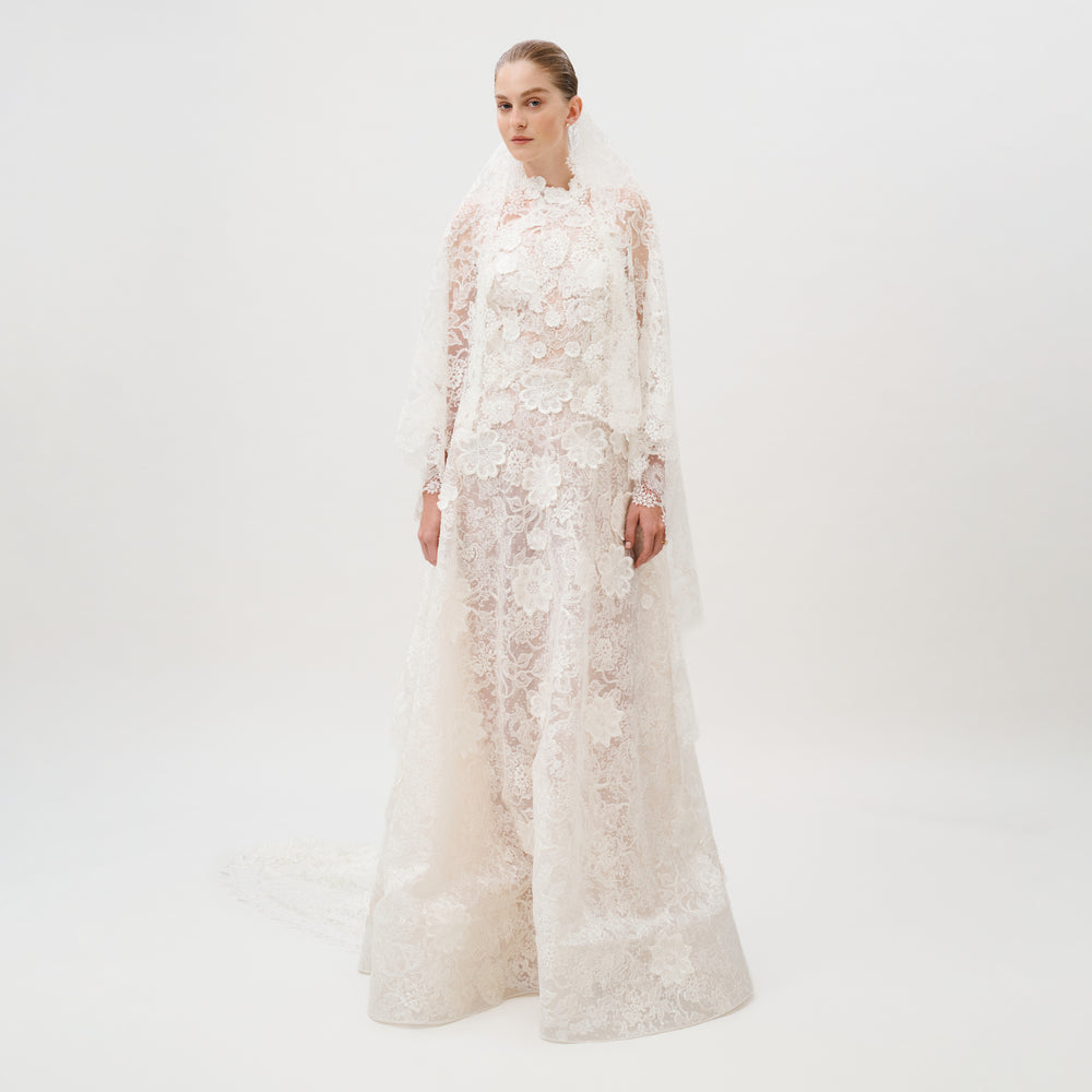 Dressed in the Danita Dress—a long, white, lace wedding gown adorned with floral patterns and crafted from exquisite Italian fabric by Colombia Atelier—a woman stands against a plain white background. Her timeless bridal look is completed with a matching lace veil.