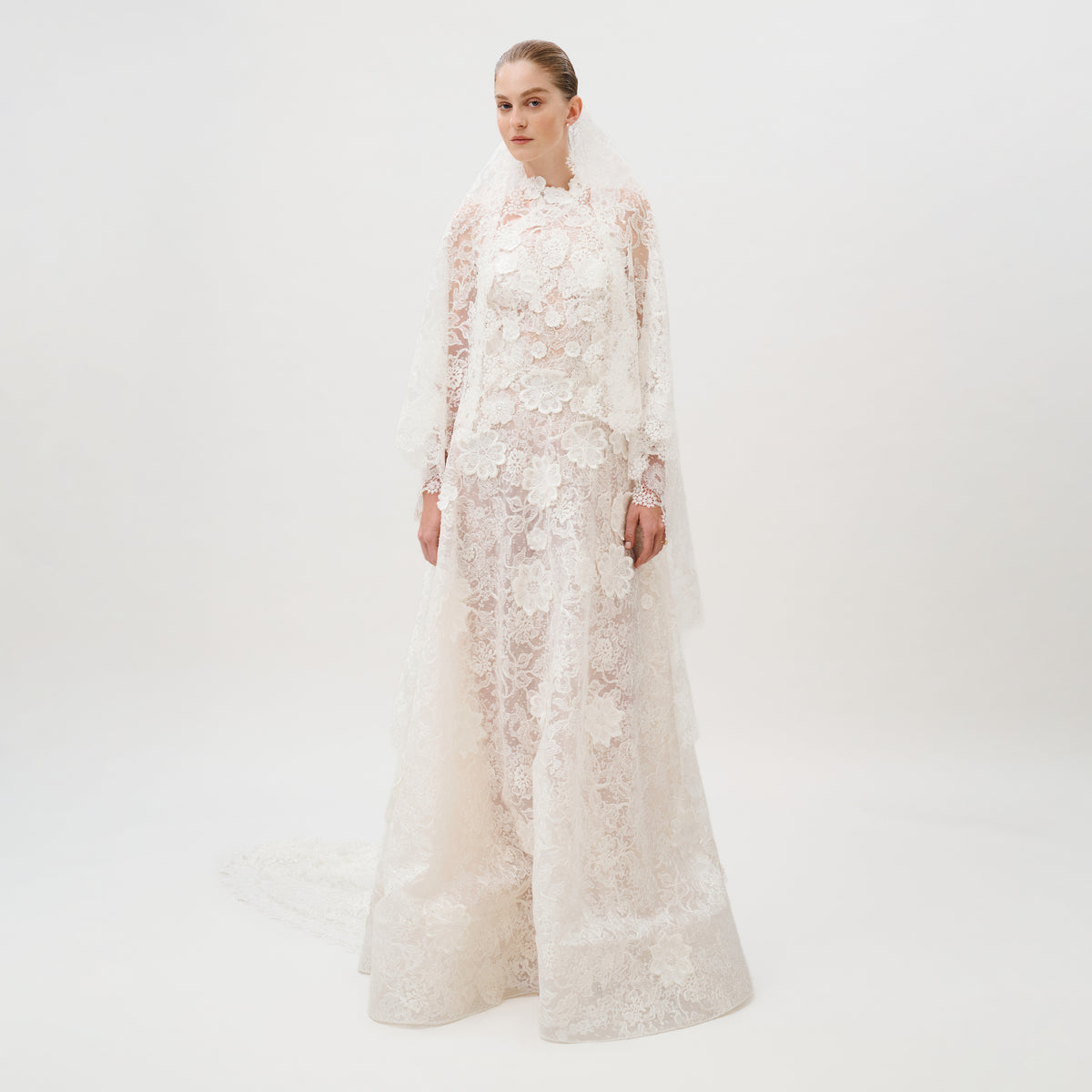 Dressed in the Danita Dress—a long, white, lace wedding gown adorned with floral patterns and crafted from exquisite Italian fabric by Colombia Atelier—a woman stands against a plain white background. Her timeless bridal look is completed with a matching lace veil.
