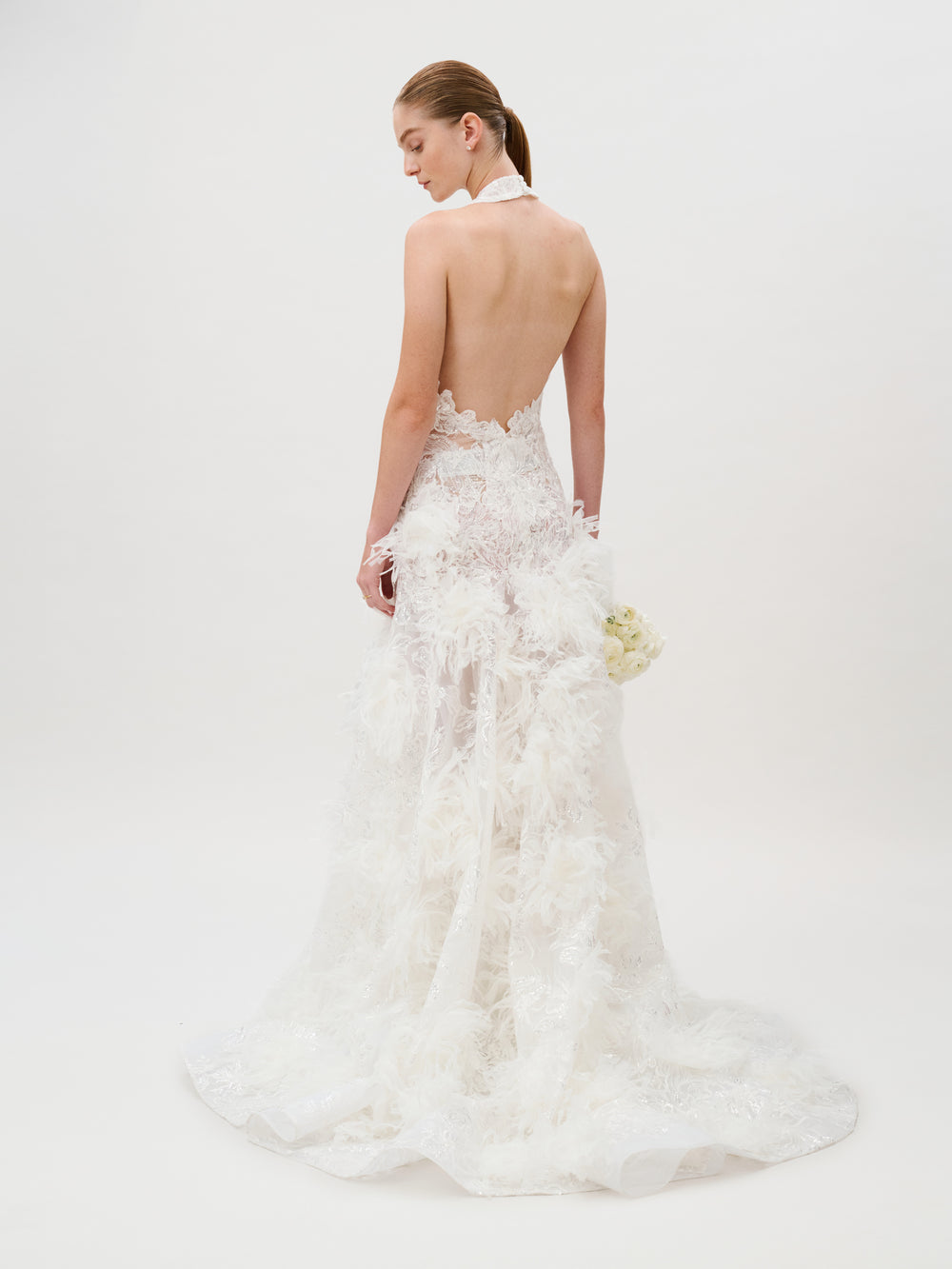 Dressed in the exquisite Darsha Dress—a backless white gown featuring lace and feather details with delicate silk trim—a woman stands holding a small bouquet of white flowers against a plain white background. This stunning dress is proudly made in Italy.