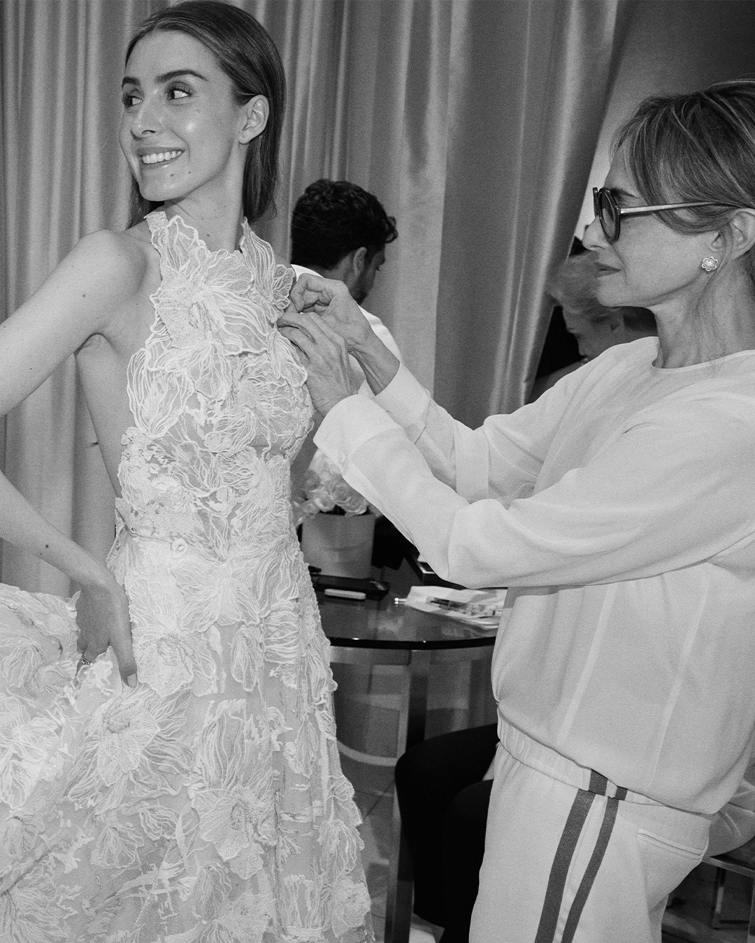 Designer adjusts a floral lace gown on a smiling model in a fitting room.