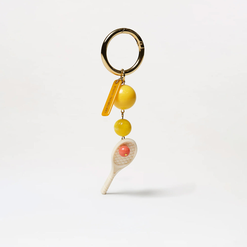 The Deuce Charm is a gold-tone metal ring keyring with a yellow tag, a large yellow bead, and a tennis racket charm featuring a small pink ball in its center, made in Colombia, shown against a plain white background.