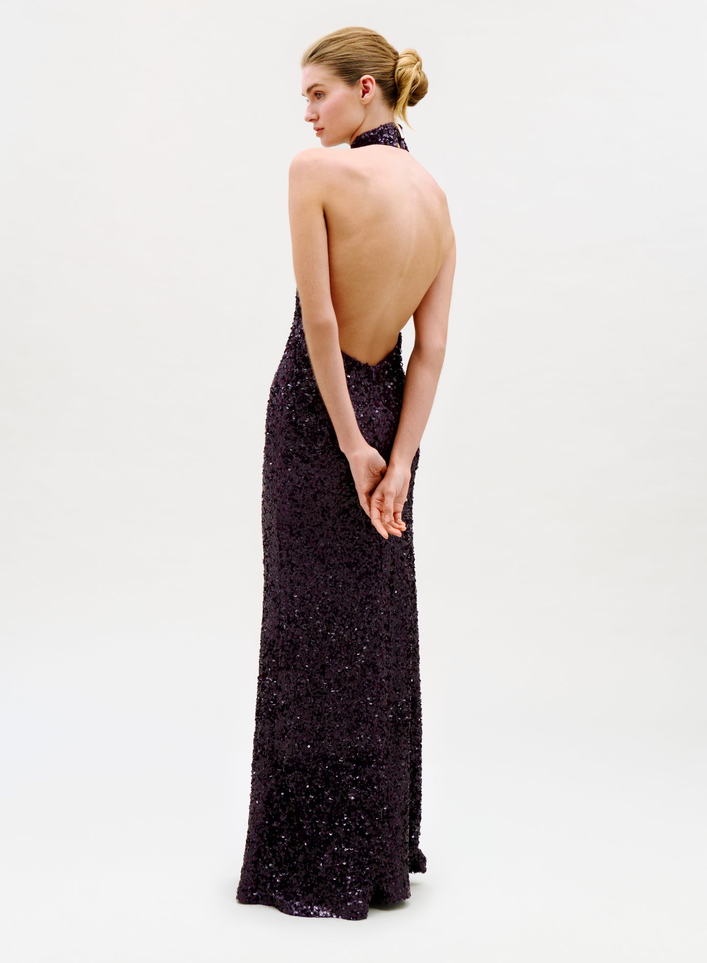 A woman in the Dido Dress—a floor-length, sleeveless, backless black sequin gown—stands with her hands clasped behind her back, looking to her left against a plain light background.