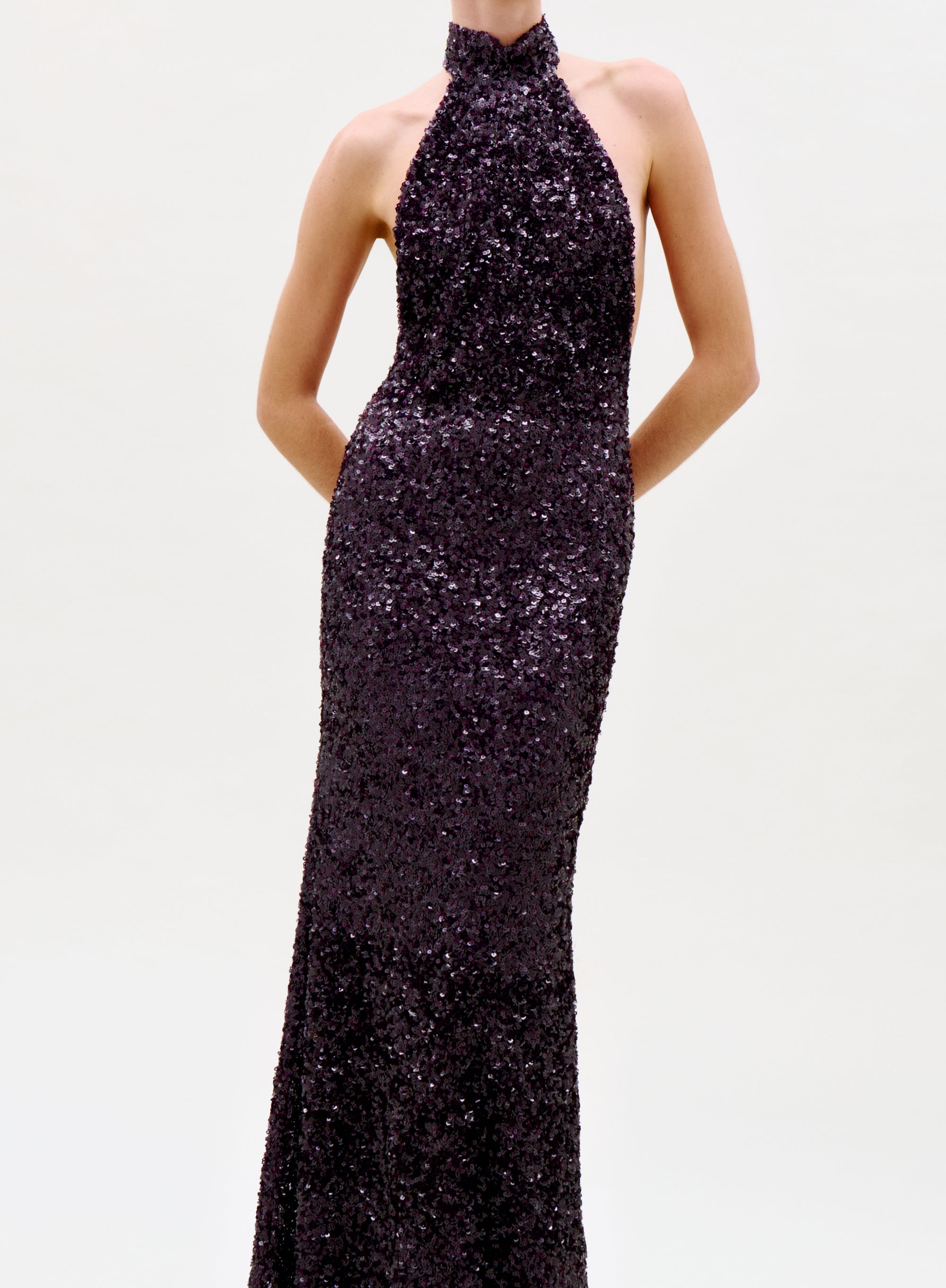 A person models the sleeveless, floor-length Dido Dress with dark sequins and a halter neck, standing hands behind their back against a plain light backdrop. The photo is cropped at the neck.