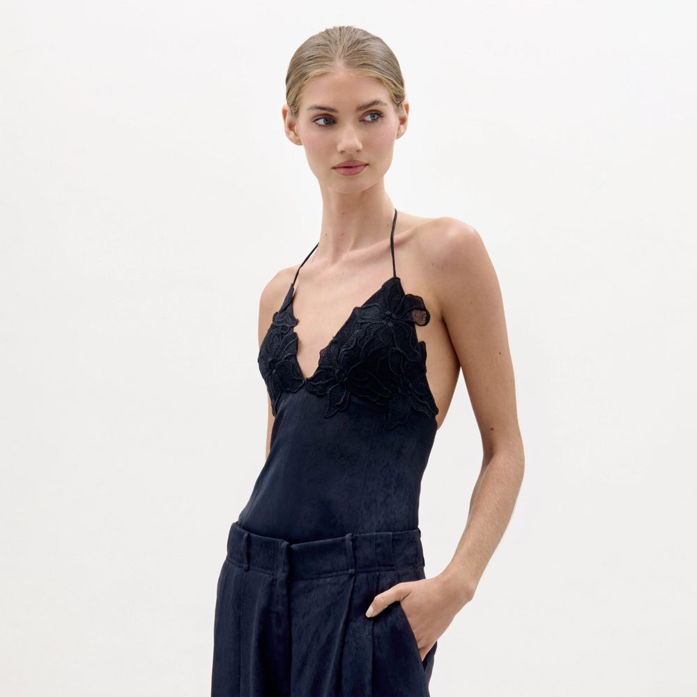 A woman with blonde hair tied back wears the Diora Top, a black halter-neck style with floral lace applique, paired with dark high-waisted pants. She poses with one hand in her pocket against a plain white background.